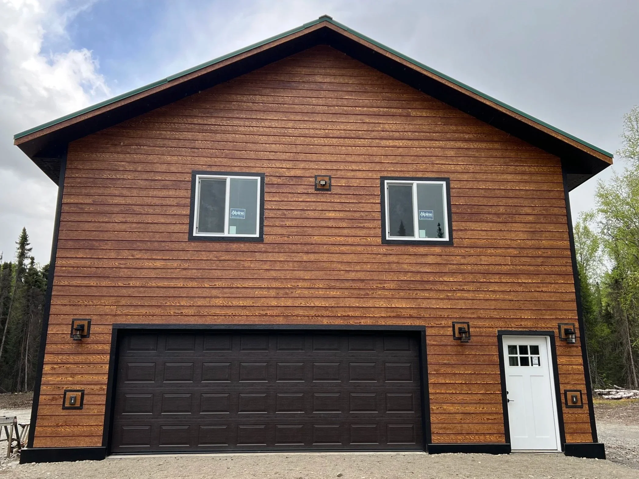 A two-story house with wooden siding, a dark garage door, a white side door, and two windows on the upper floor, surrounded by greenery and a partly cloudy sky.