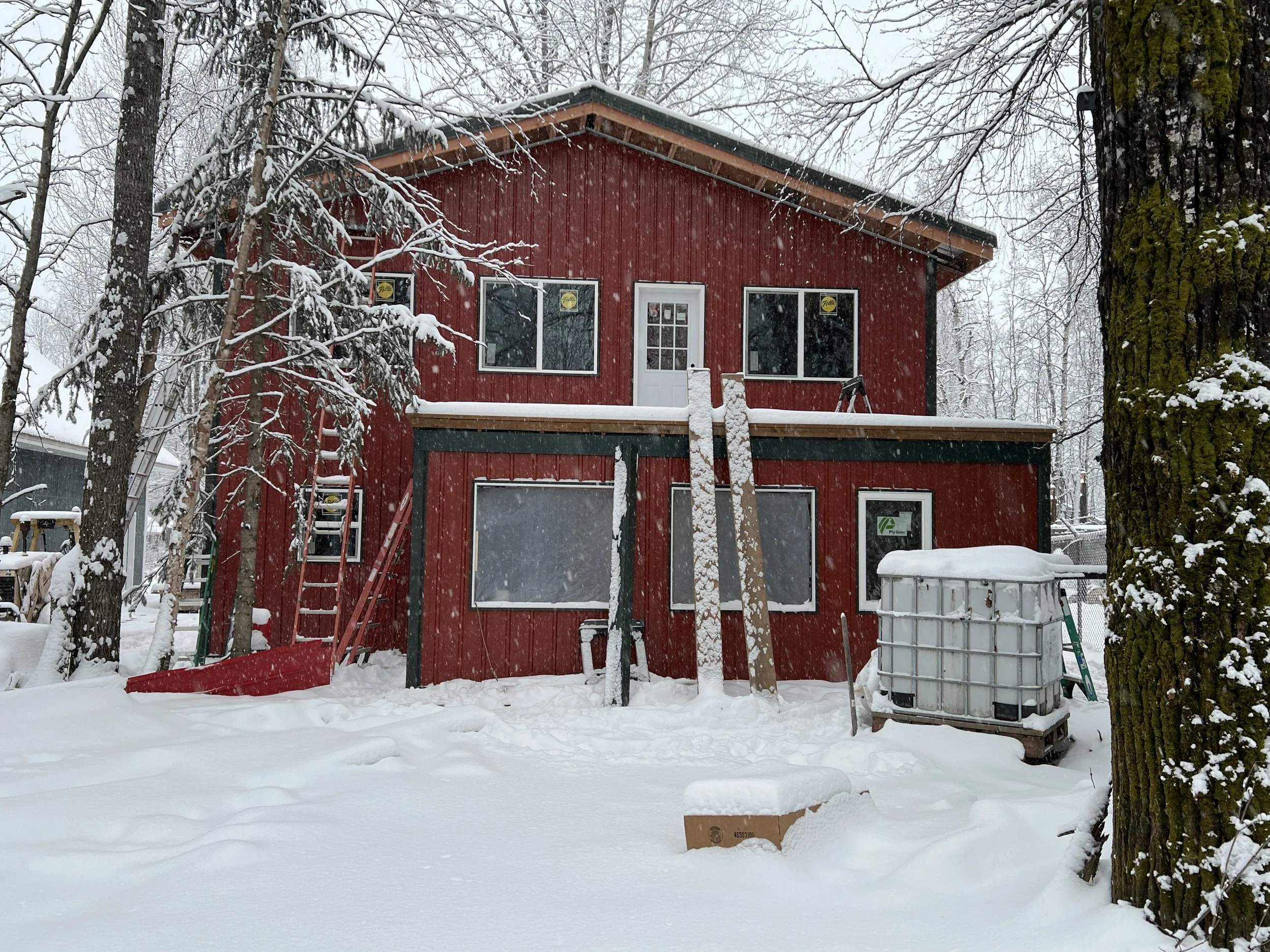 A two-story red house with a green roof and white window frames is under construction in a snowy environment. There are ladders leaning against the house, with snow on them, and a large container filled with snow nearby. Snow-covered trees and a cloudy sky are visible in the background.