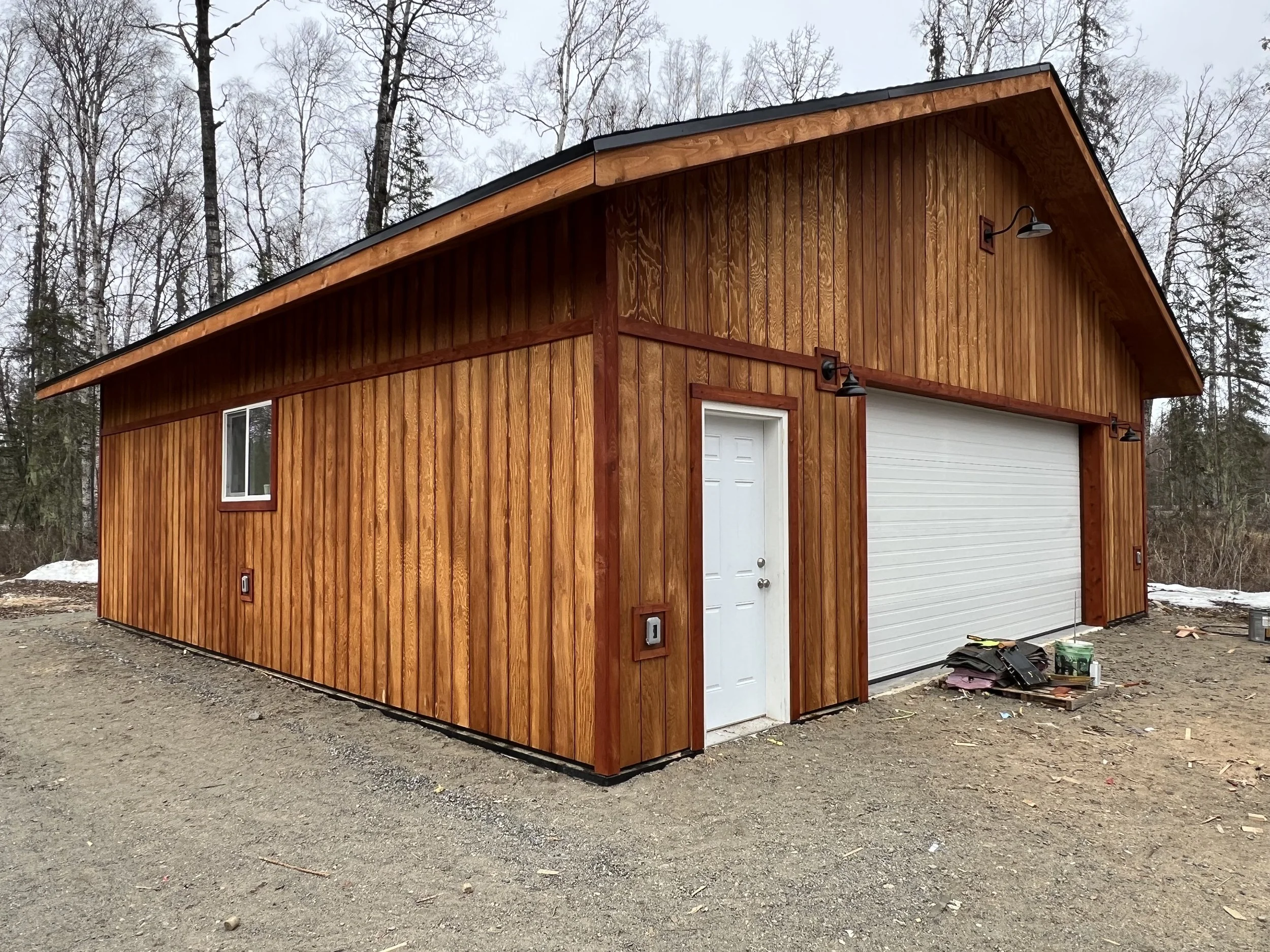 A wooden garage with a white door and a garage door, situated on a dirt lot with trees in the background.
