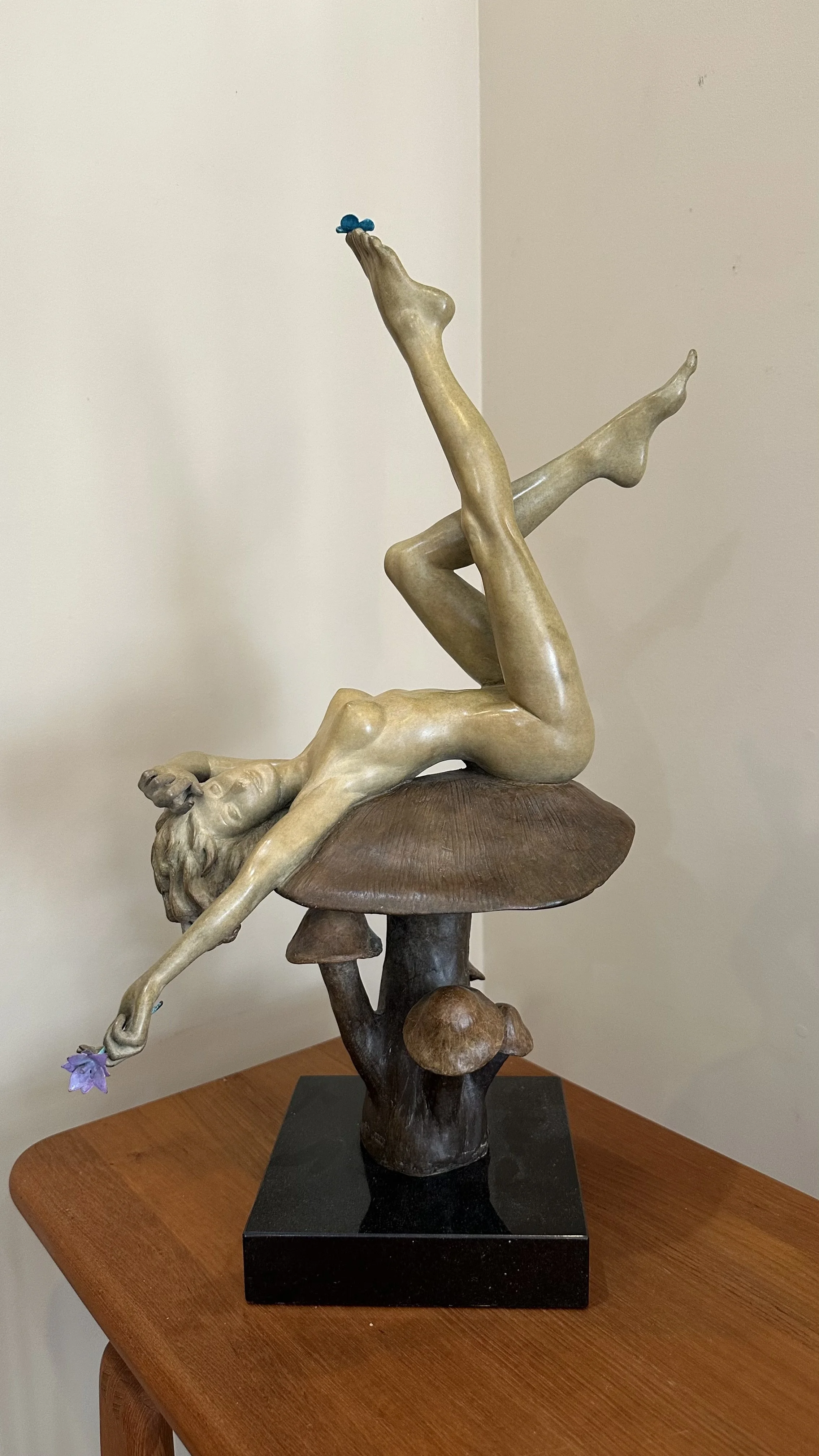 Finally Spring by Danielle Anjou, cast bronze figurative sculpture of a reclining nude woman atop a mushroom, holding a flower with legs extended playfully upward.