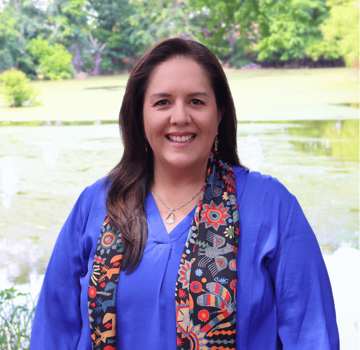 Image of Maria Fernandez wearing a cobalt blue blouse and decorative Mexican, long brown hair smiling, with a pond behind her.