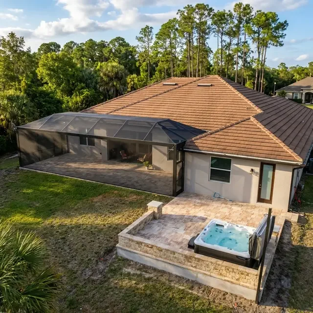 Backyard of a house with a screened patio, paver patio, small hot tub, and surrounding green lawn and trees.