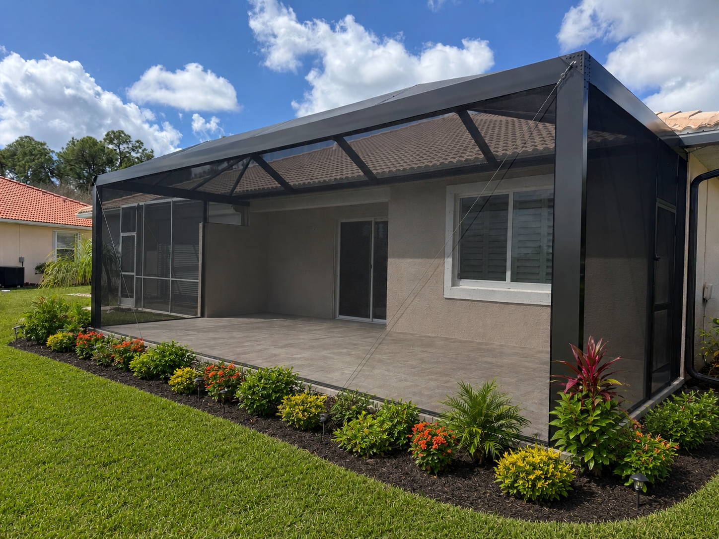Backyard patio with a screened enclosure, surrounded by a well-maintained lawn and flower bed, on a sunny day with partly cloudy sky.