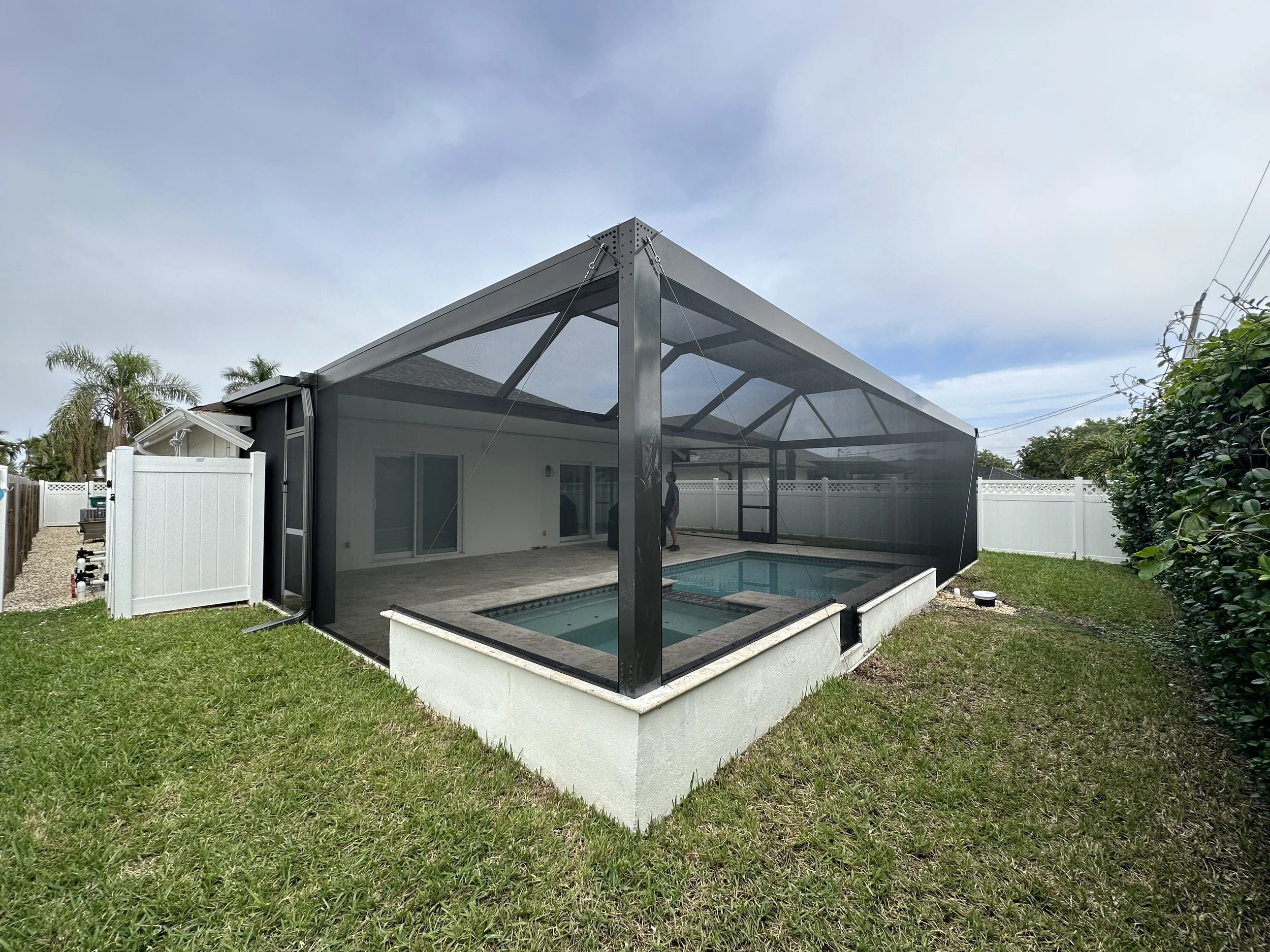 A backyard with a screened-in porch, pool, grass, and a white fence. There are palm trees and utility poles in the background under a partly cloudy sky.