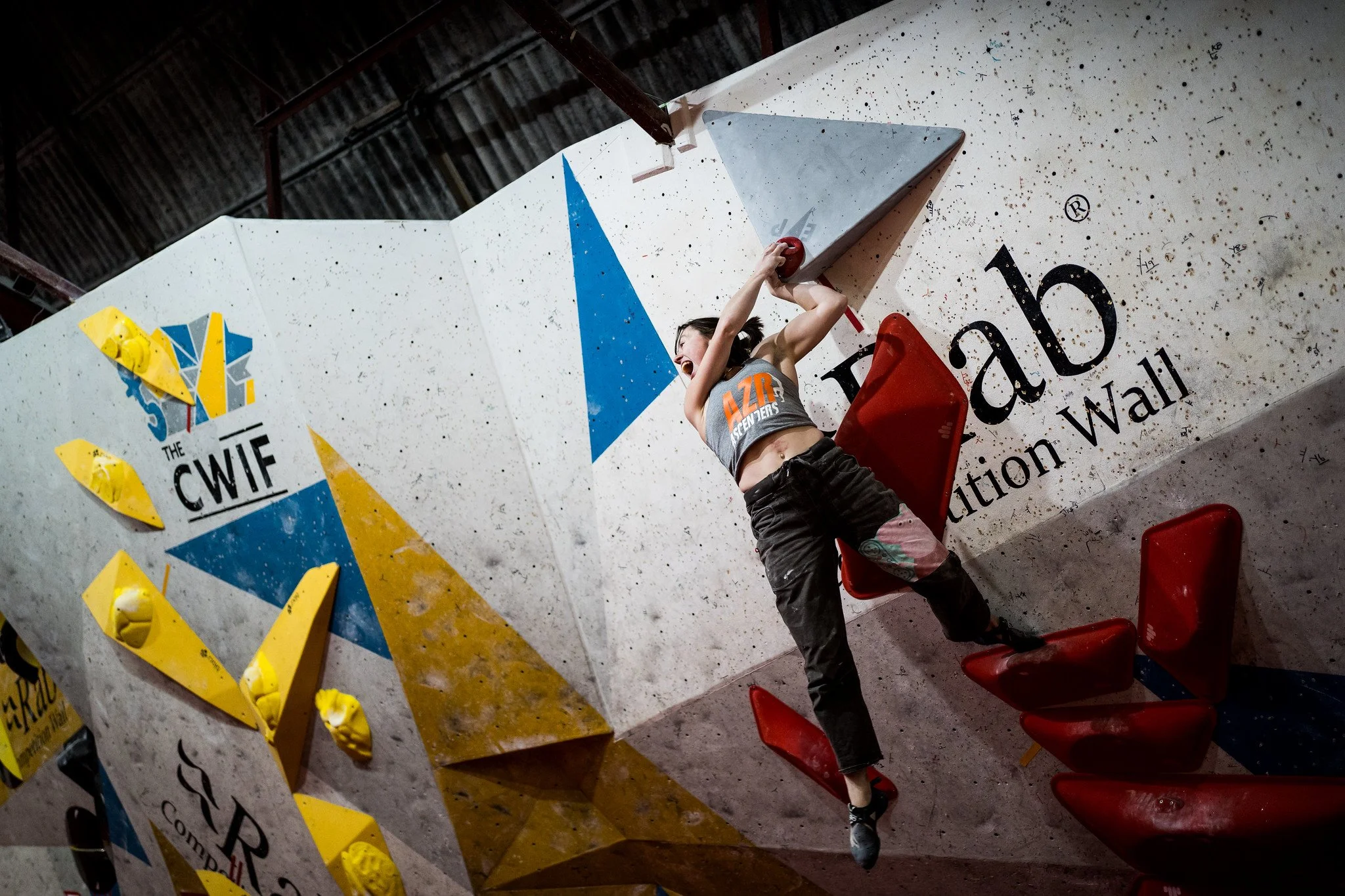 Female rock climber on an indoor climbing wall, reaching for holds with a look of concentration. She is wearing a gray sports top and black pants, with a red crash pad below her. The climbing wall is decorated with various colored holds and signage.
