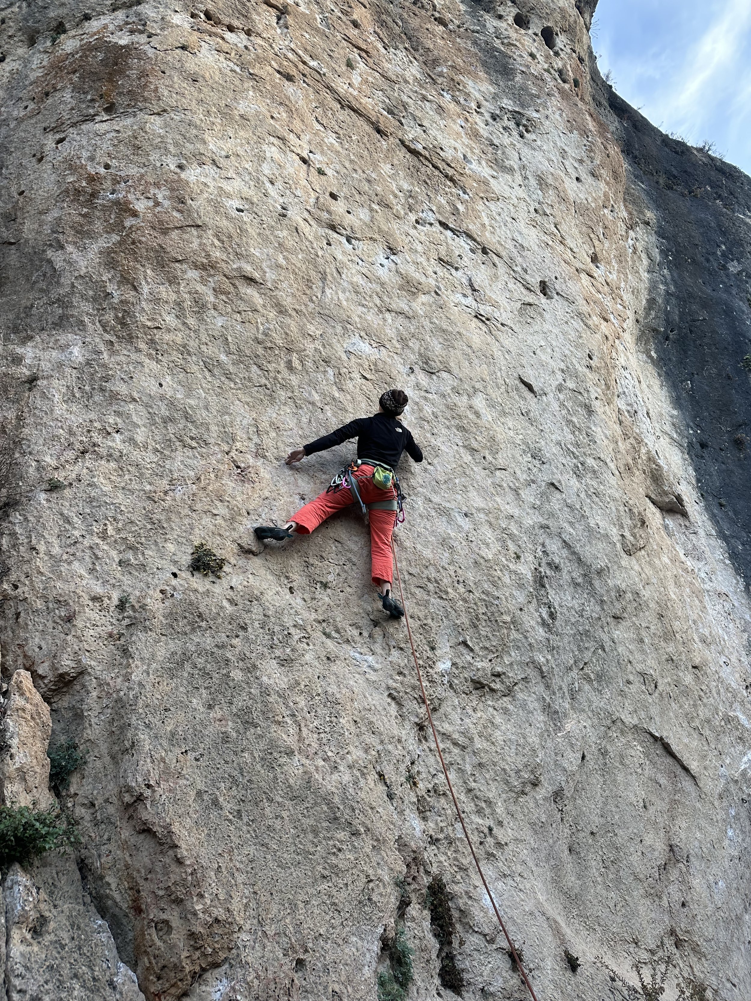 A person rock climbing on a steep, rugged cliff face with a safety rope attached, wearing a black jacket, red pants, and climbing gear.