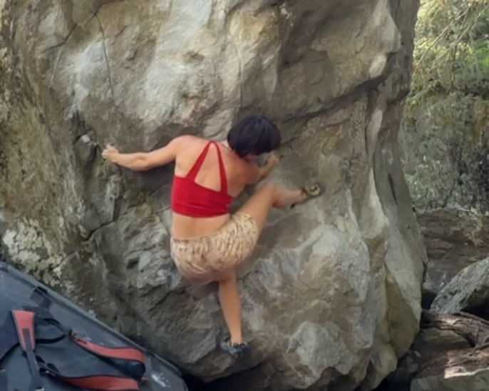Person wearing a red sports bra and patterned shorts rock climbing on a large boulder outdoors.