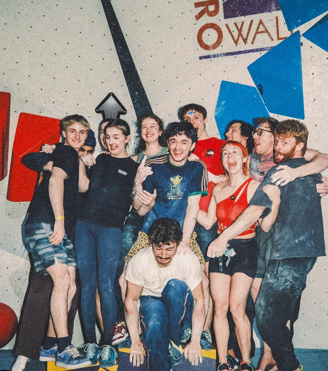 Group of people celebrating after indoor rock climbing, with many smiling and cheering, in front of climbing wall featuring colorful holds and abstract designs.