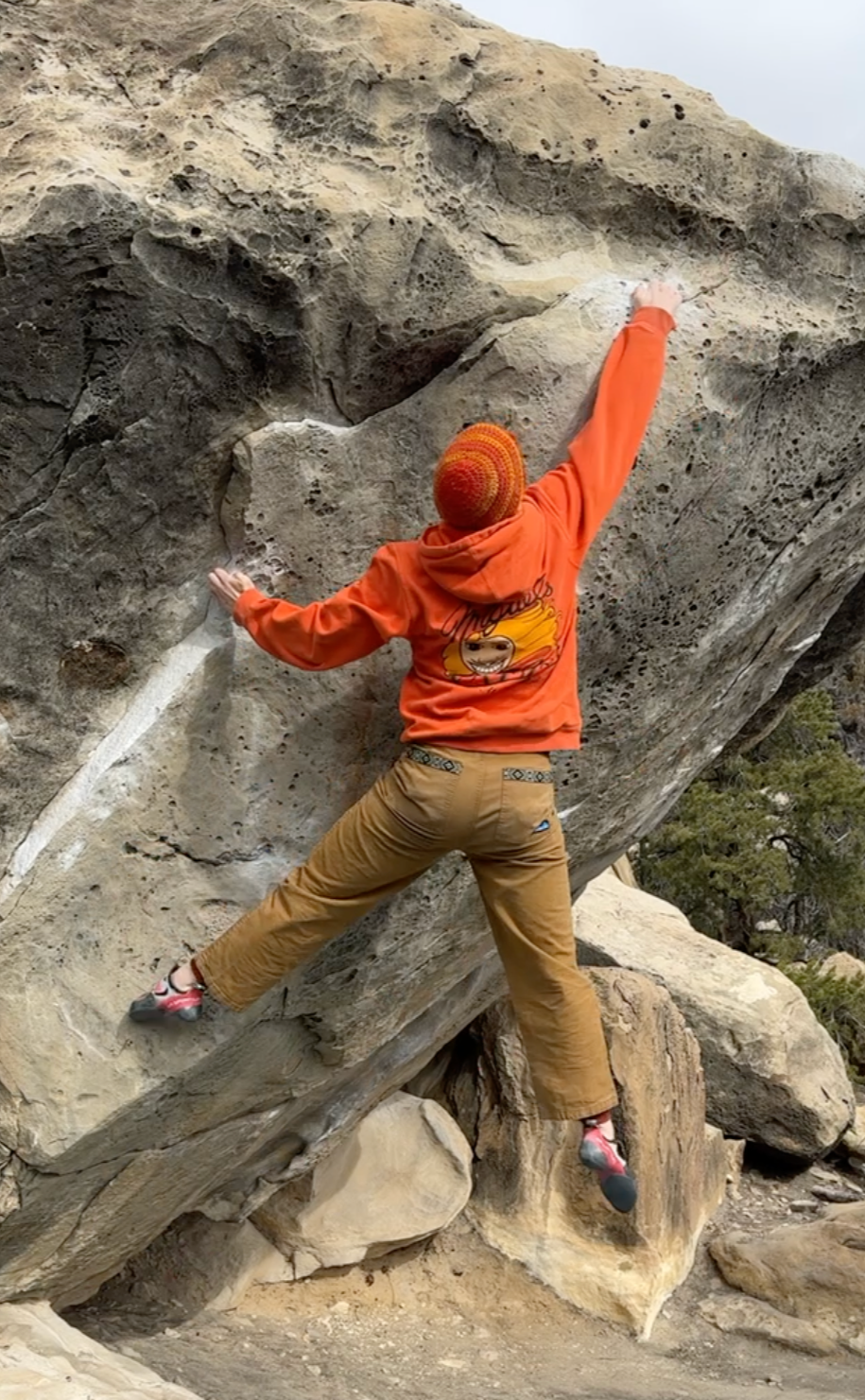 Person wearing an orange hoodie and red beanie climbing a large boulder outdoors.