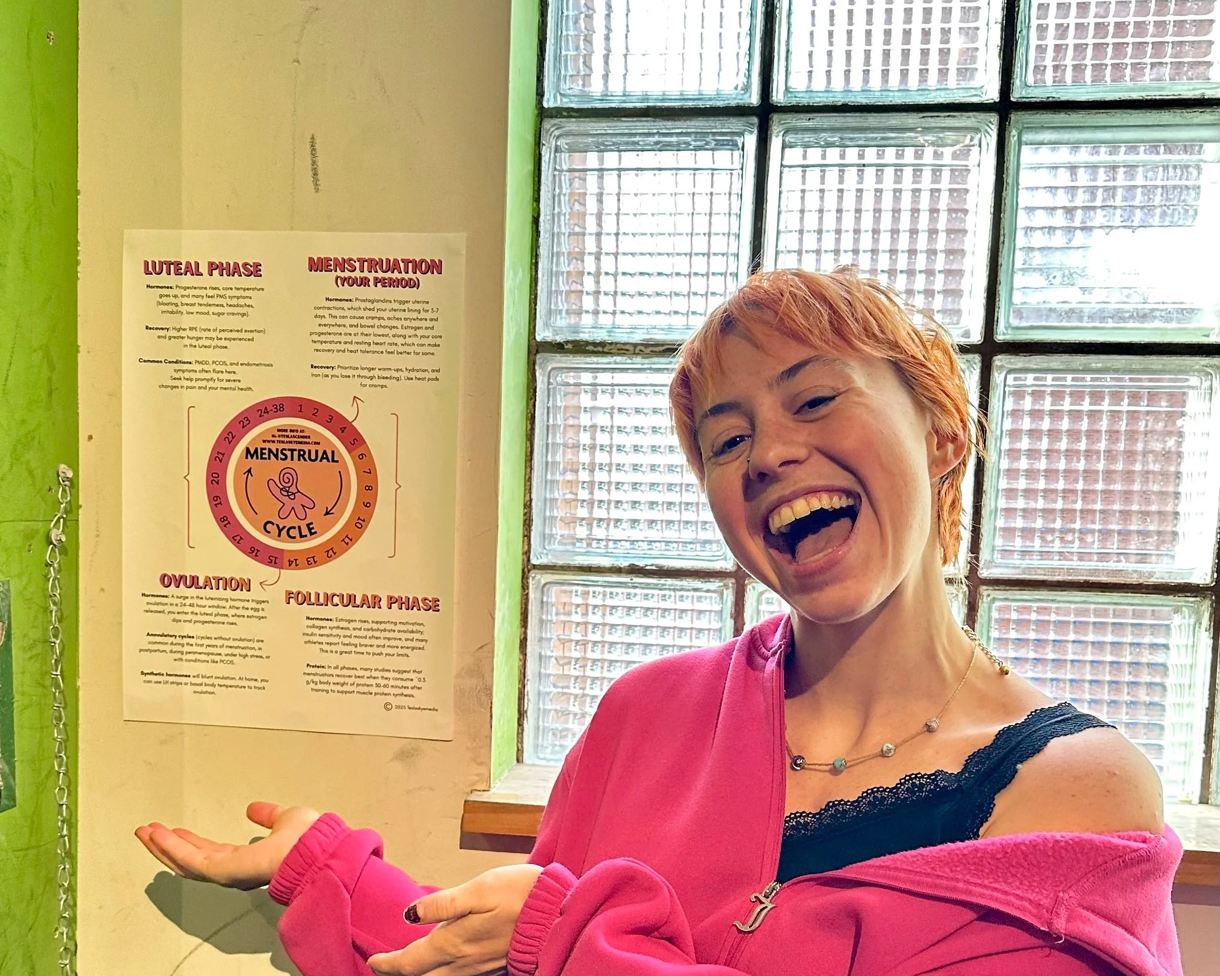 A woman with red hair, wearing a pink hoodie, smiling and gesturing with her hands while standing in front of a wall with educational posters about the menstrual cycle and phases.
