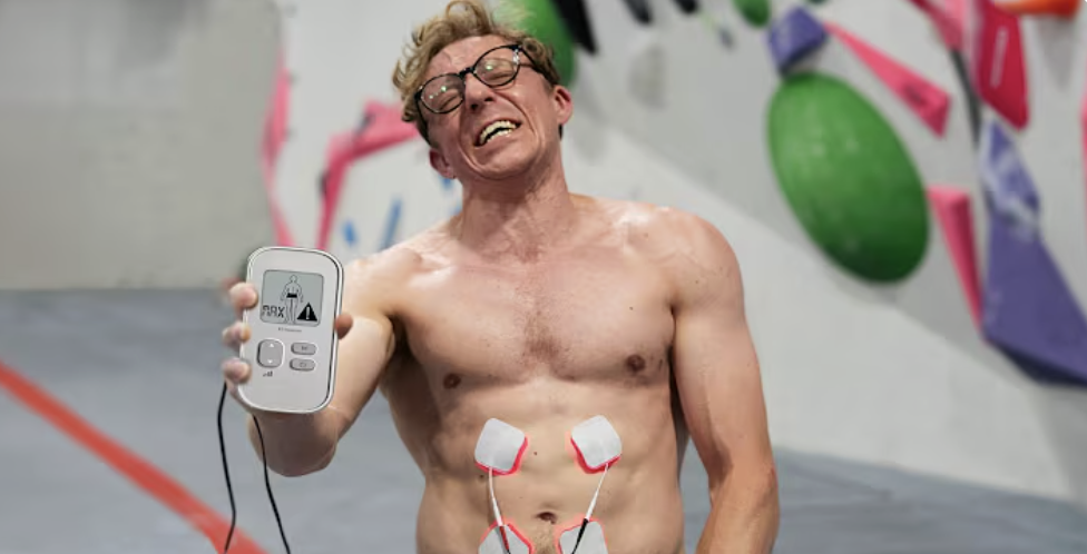 A shirtless man with glasses smiling and holding a medical device in his hand, standing in an indoor climbing gym with colorful climbing holds on the wall.