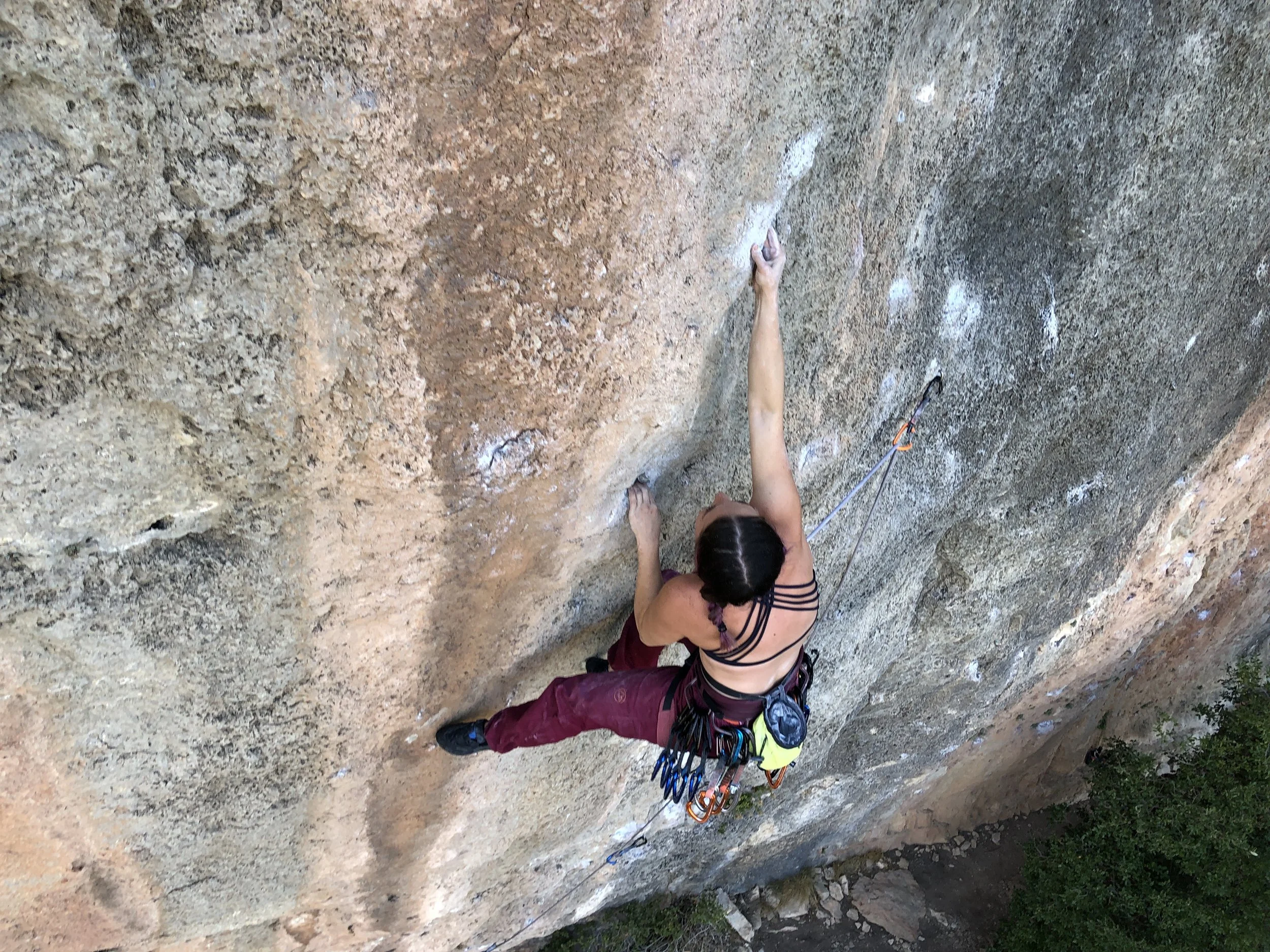 A woman with dark hair in a braid, wearing a harness and climbing shoes, is in the middle of climbing an indoor rock wall with various holds.
