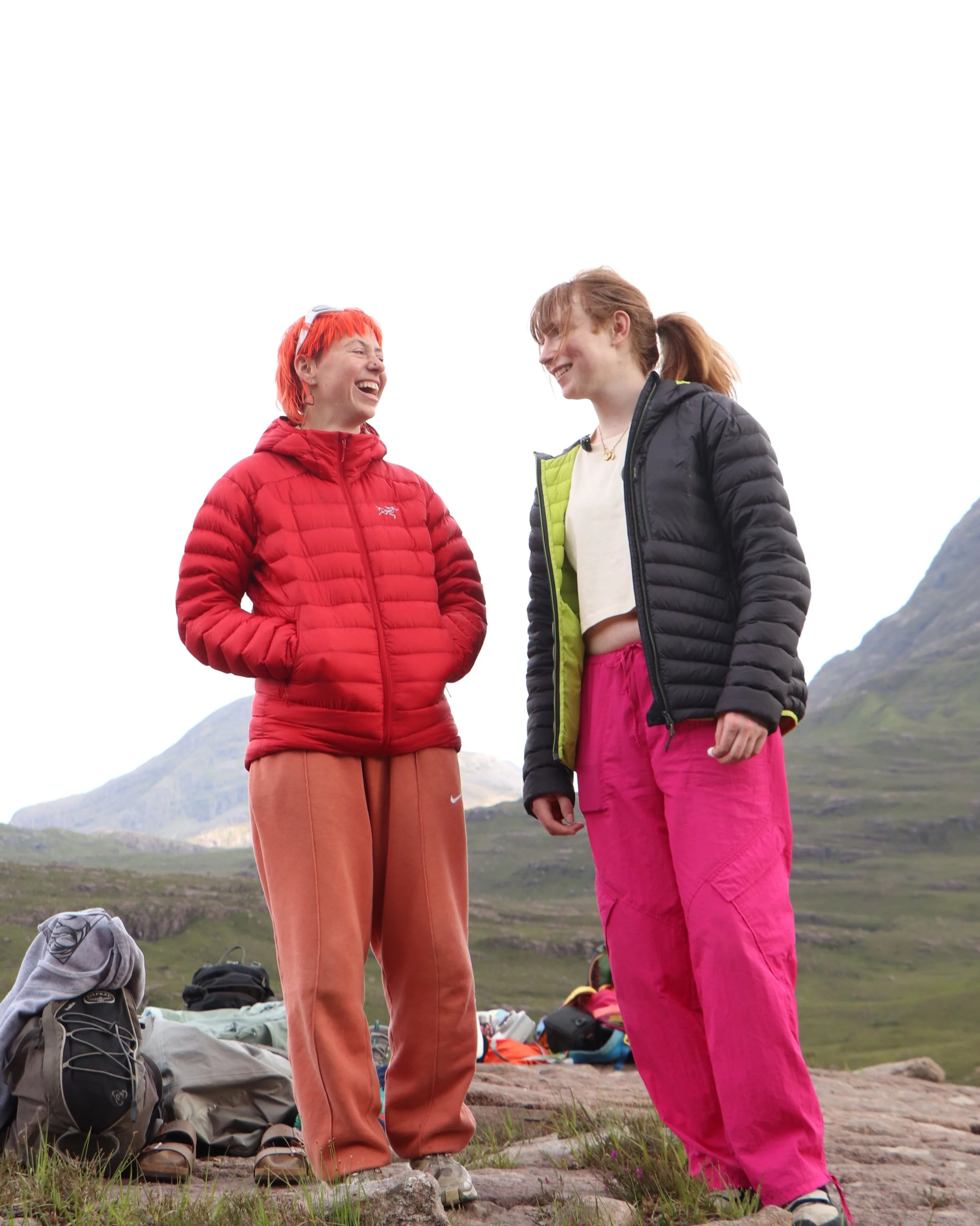Two young women wearing colorful outdoor jackets are standing outdoors on a grassy area, smiling and talking to each other. There are backpacks and outdoor gear on the ground in front of them, with mountains and cloudy sky in the background.