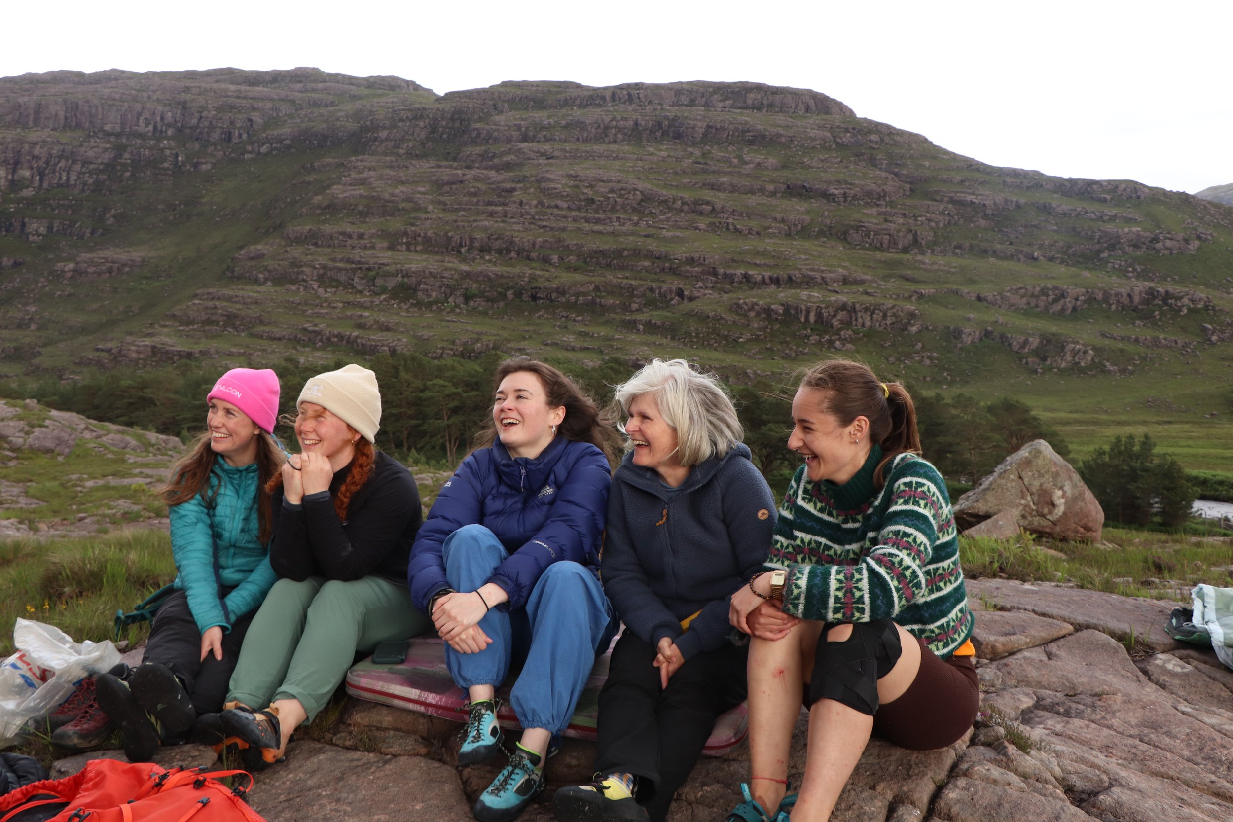 Group of five women sitting outdoors on rocks in front of a mountain landscape, laughing and enjoying a hike.
