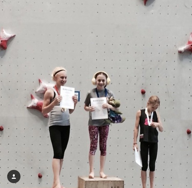 Three girls standing on a podium during a rock climbing competition, holding certificates and medals. They are in front of a climbing wall with pink star-shaped holds.