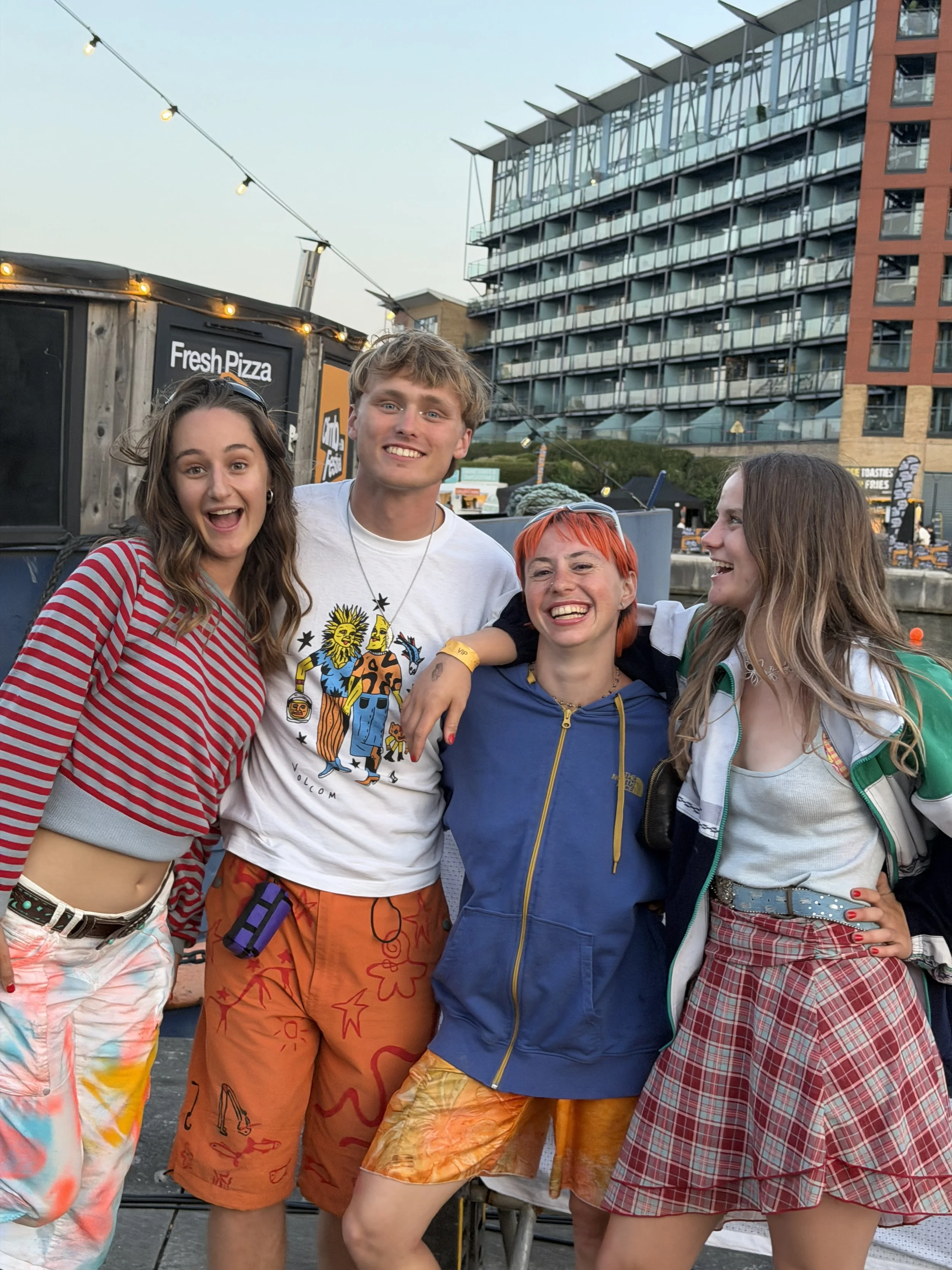 Group of five smiling young adults at an outdoor event during daytime, standing close together, wearing casual and colorful summer clothes, with a modern building and a food stand advertising pizza in the background.