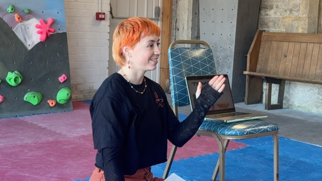 A woman with short orange hair, wearing a black shirt and necklace, smiling and gesturing with her hand while sitting in an indoor climbing gym with a rock climbing wall in the background.