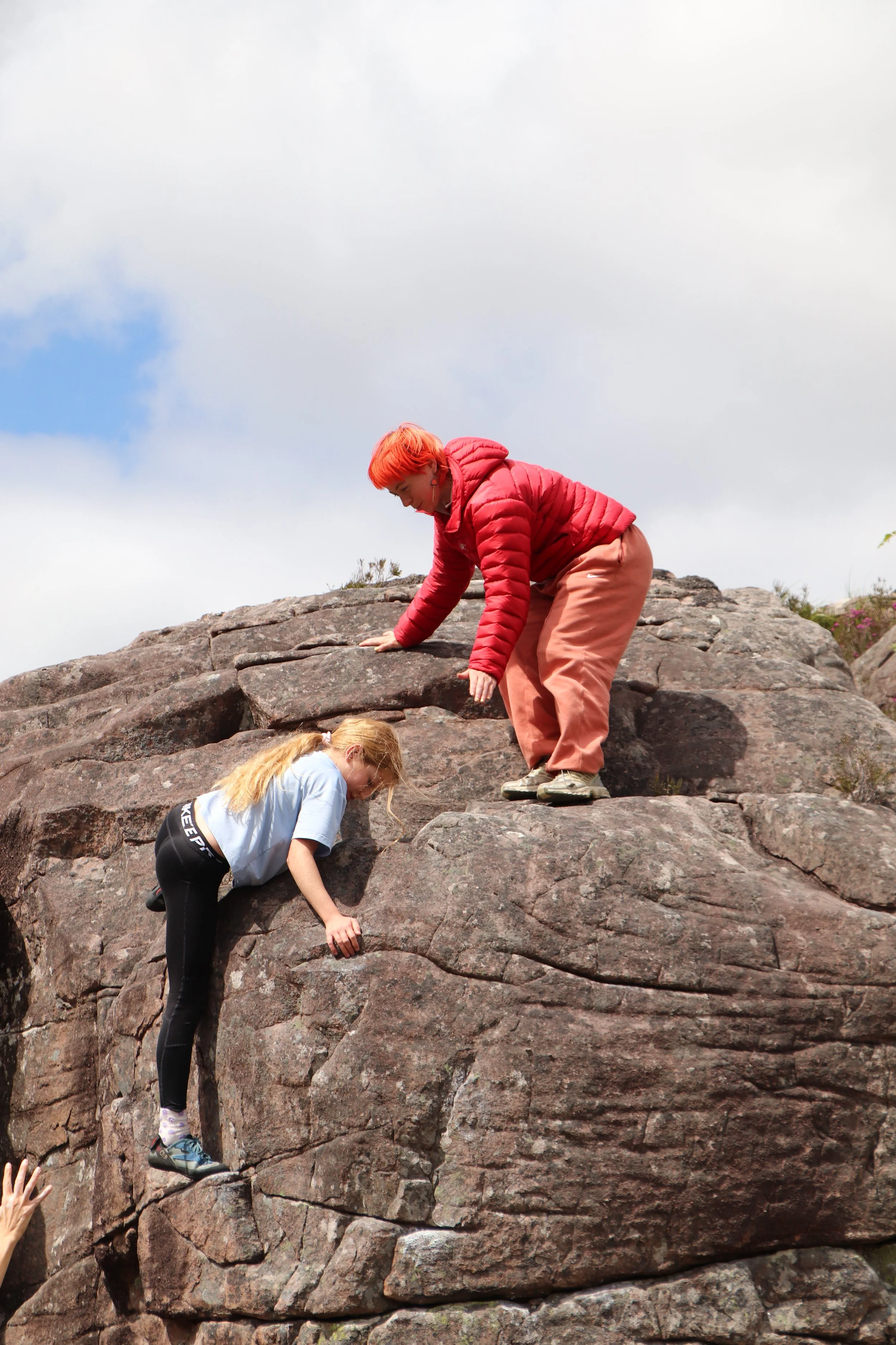 Two children climbing a large rock formation outdoors in a natural setting with cloudy sky above.