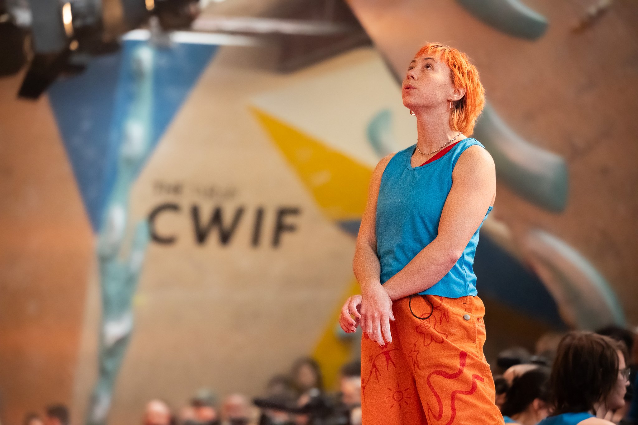A woman with short orange hair and hoop earrings is wearing a blue tank top and orange pants with red designs, standing in front of an indoor rock climbing wall at the competition.