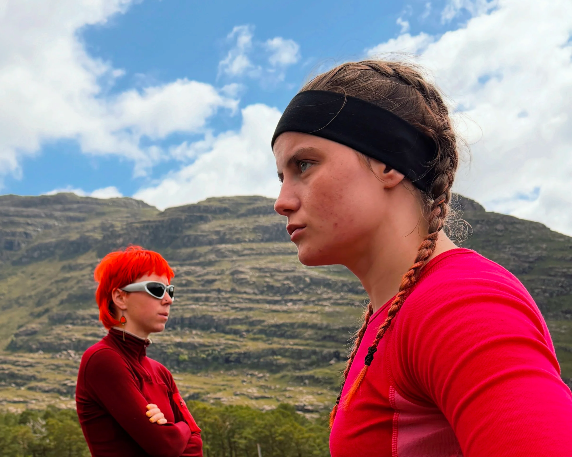 Two women outdoors with mountains and cloudy sky in the background. One woman with braided hair and a black headband in the foreground, wearing a red top. The other woman with short bright red hair, sporting sunglasses and a red top in the background.