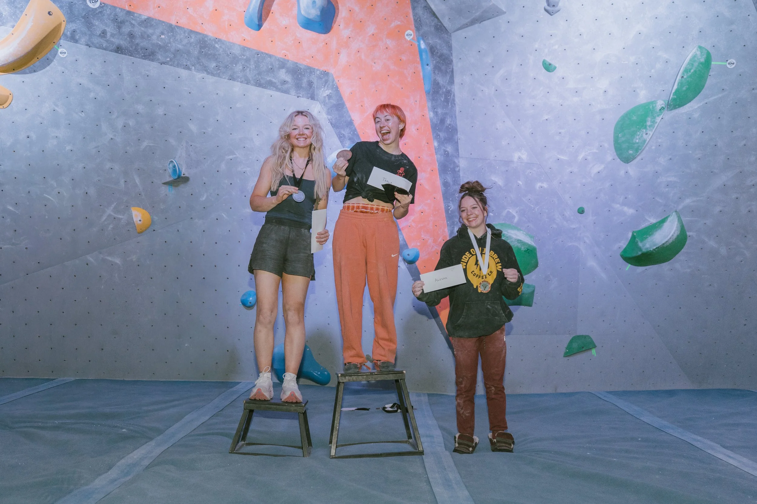 Three young women standing on a climbing competition awards platform, holding medals and certificates, with a colorful indoor climbing wall in the background.