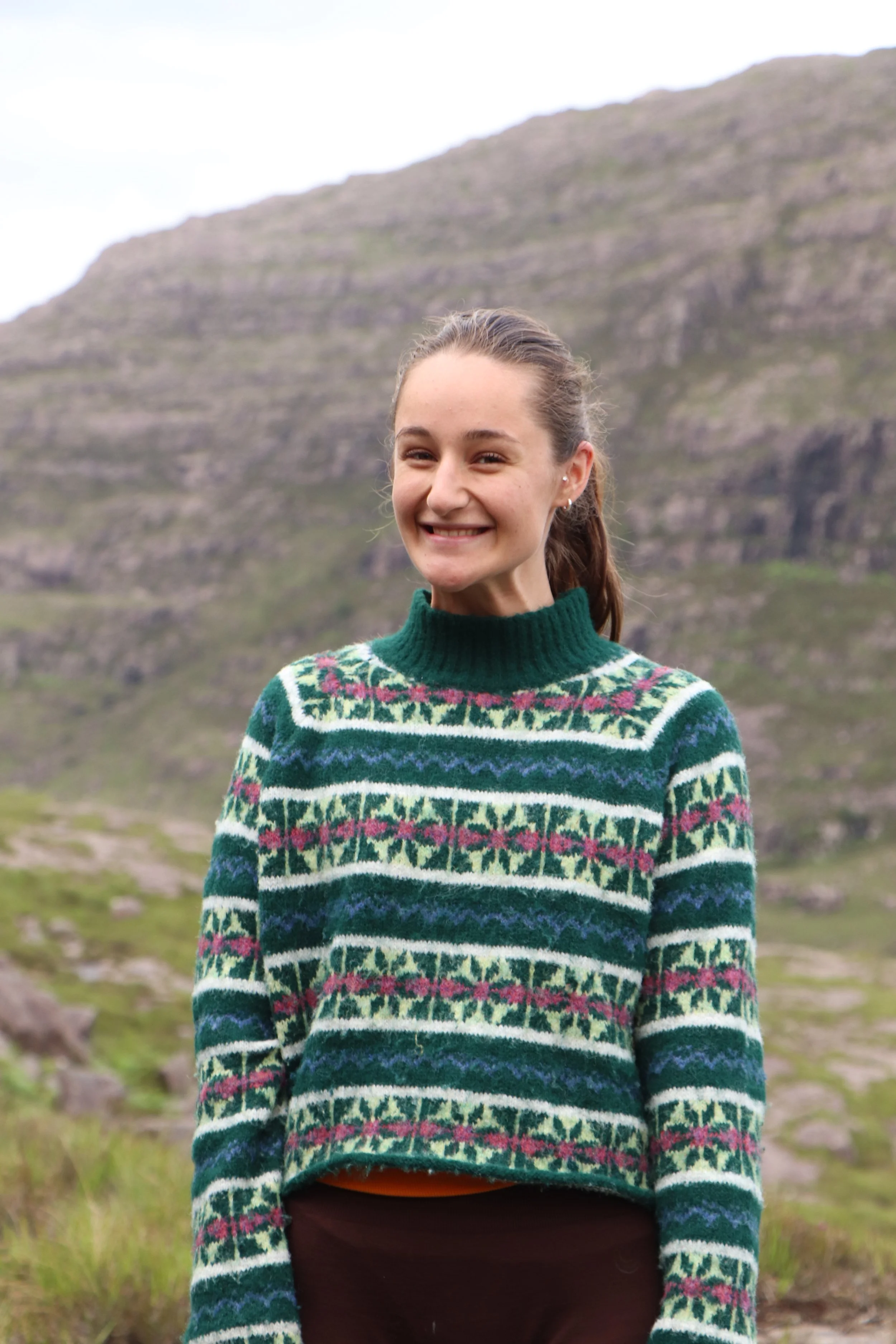 A woman smiling outdoors with mountains in the background, wearing a green, pink, purple, white, and navy striped patterned sweater.