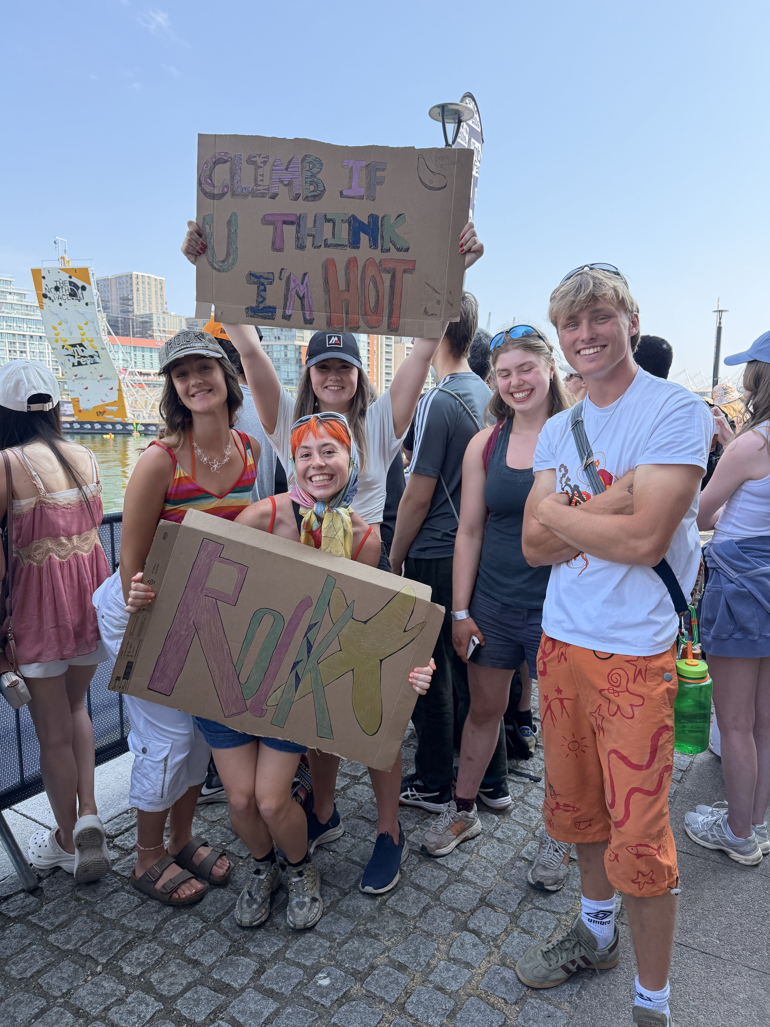 Group of young people smiling at a waterfront event, holding handmade signs with colorful doodles and text, with city buildings and boats in the background.