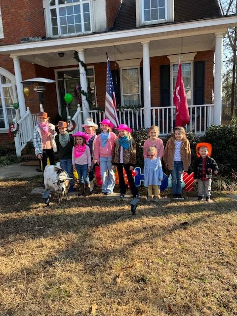 Children dressed in pink, cowboy hats, and casual clothes standing in front of a house with American and Alabama flags, along with an adult and a goat.