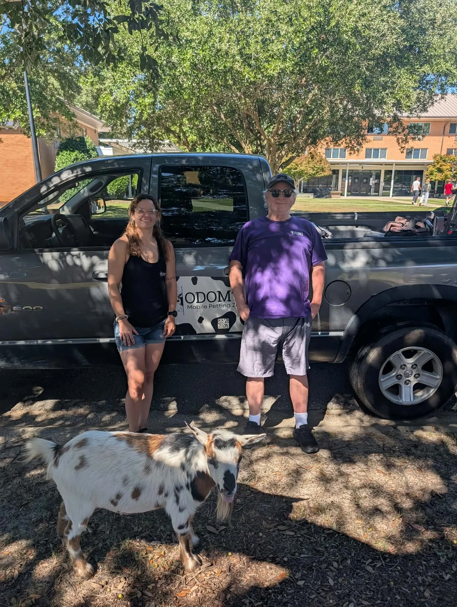 Two people standing in front of a black truck with a 'GODOM' logo, a dog with black and white spots, trees providing shade, and a building in the background on a sunny day.