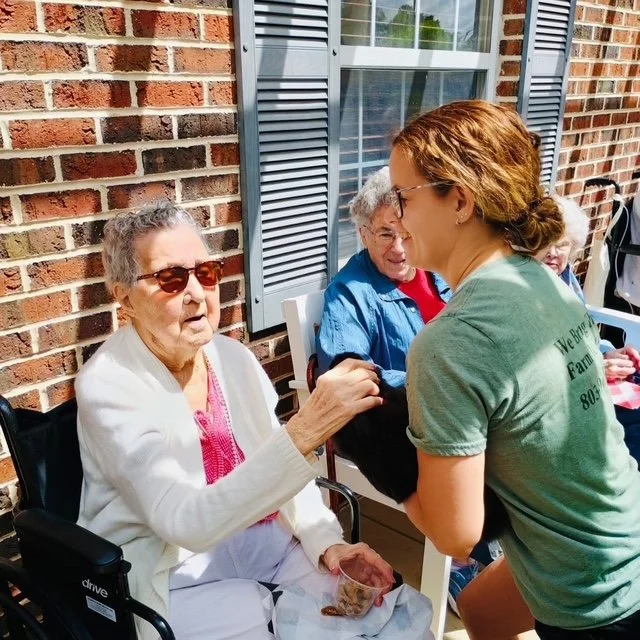 A young woman in a green T-shirt talking to an elderly woman in a wheelchair outside a brick building with blue shutters.