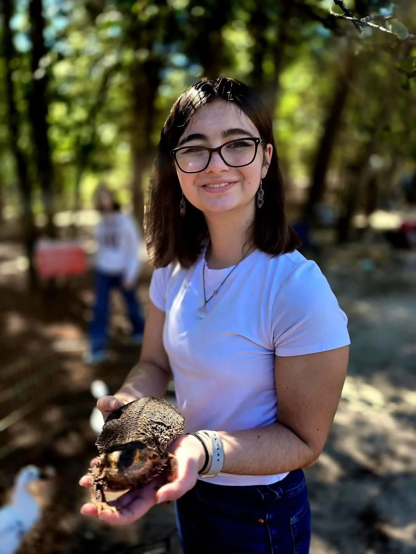 A woman with glasses and earrings holding a small bird in her hands outdoors. She is smiling and wearing a white t-shirt. The background shows blurred trees and a person in the distance.