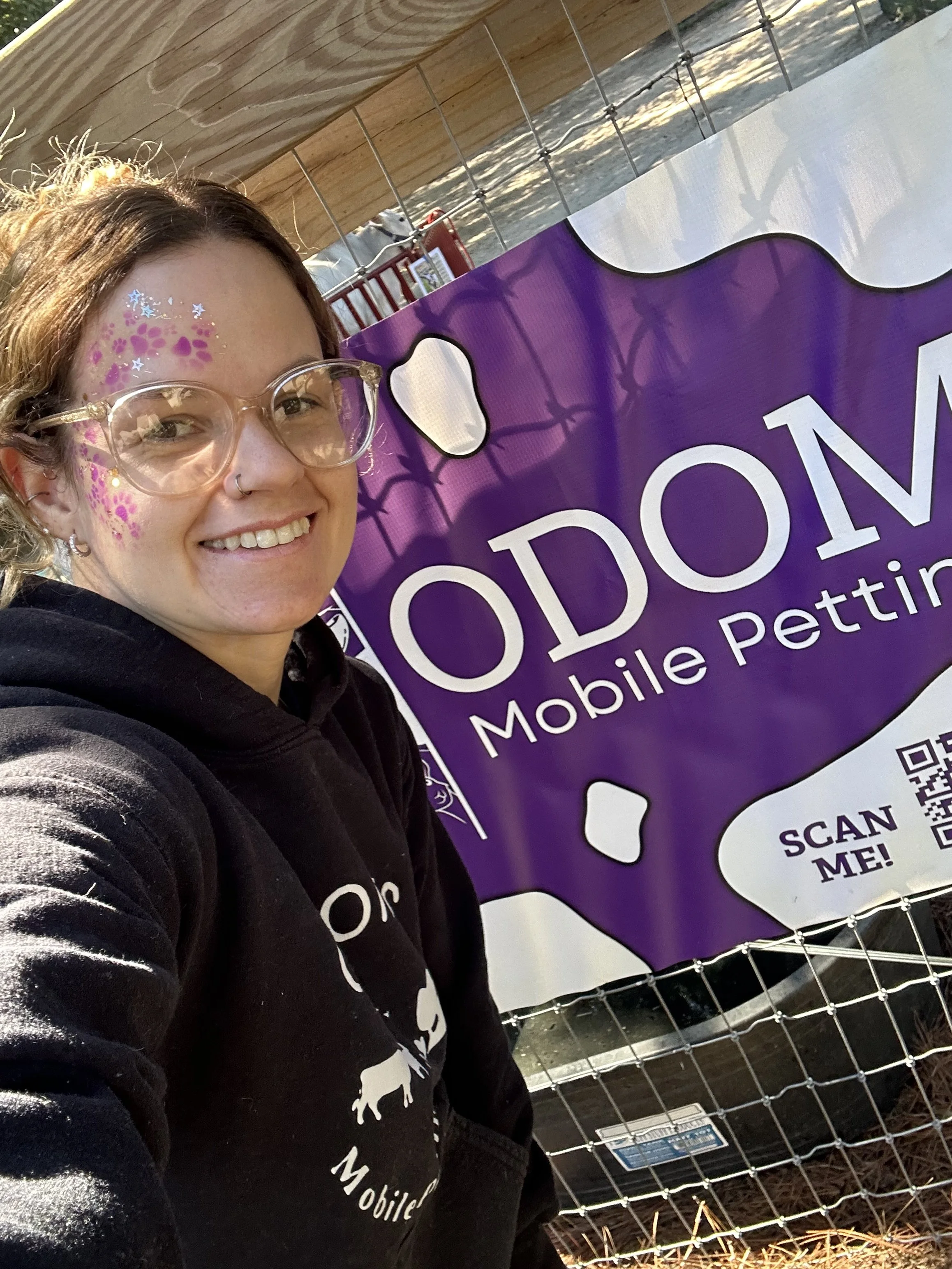 A woman with glasses smiling near a purple sign that reads 'PODON Mobile Petting' with a drawing of a dog and a speech bubble saying 'SCAN ME!'