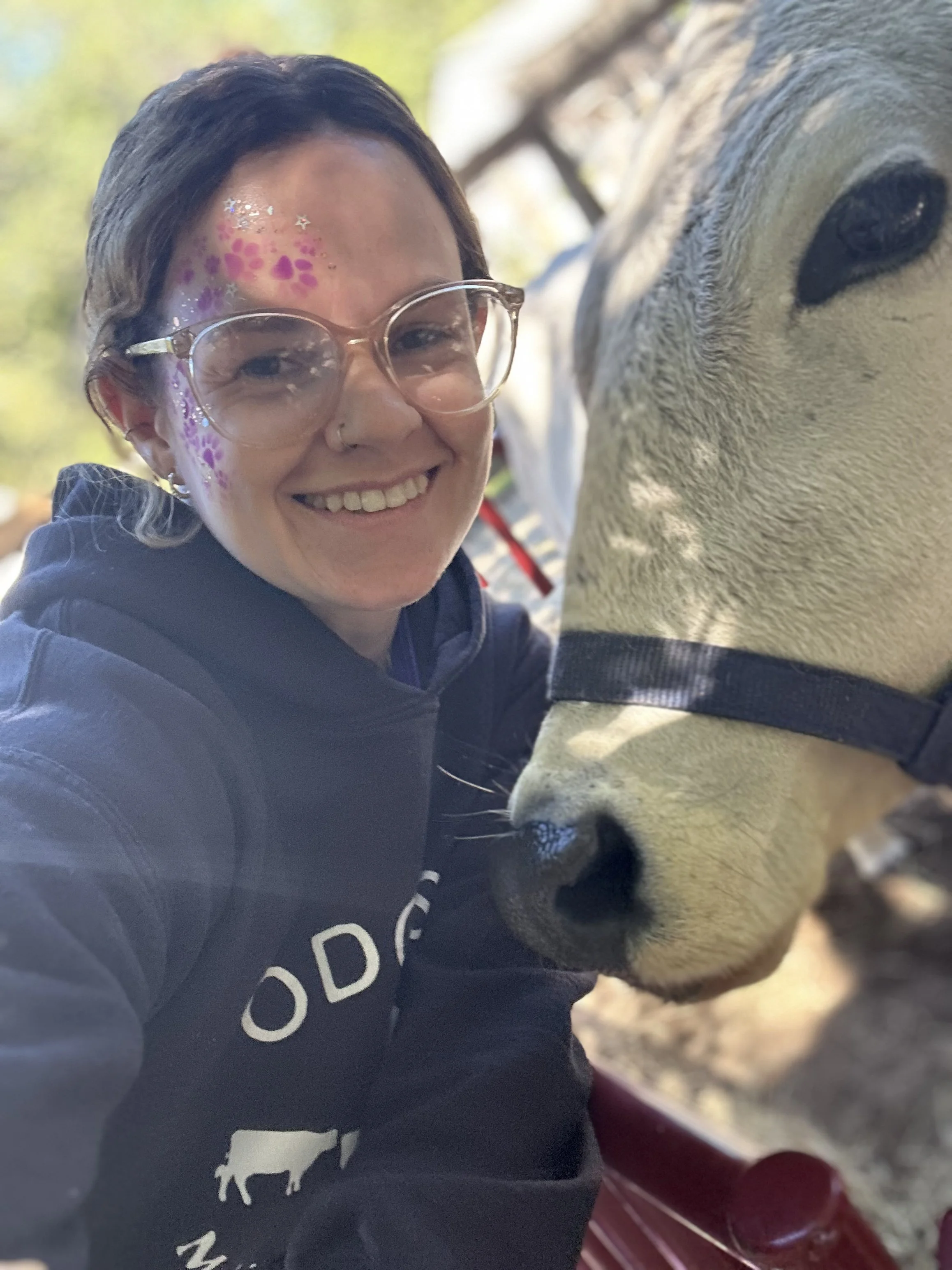 A smiling woman with glasses and face paint taking a selfie with a dog outdoors, with trees in the background.
