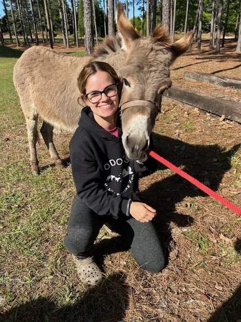 A woman with glasses kneels next to a donkey in a wooded outdoor setting, smiling at the camera, holding a red leash attached to the donkey.