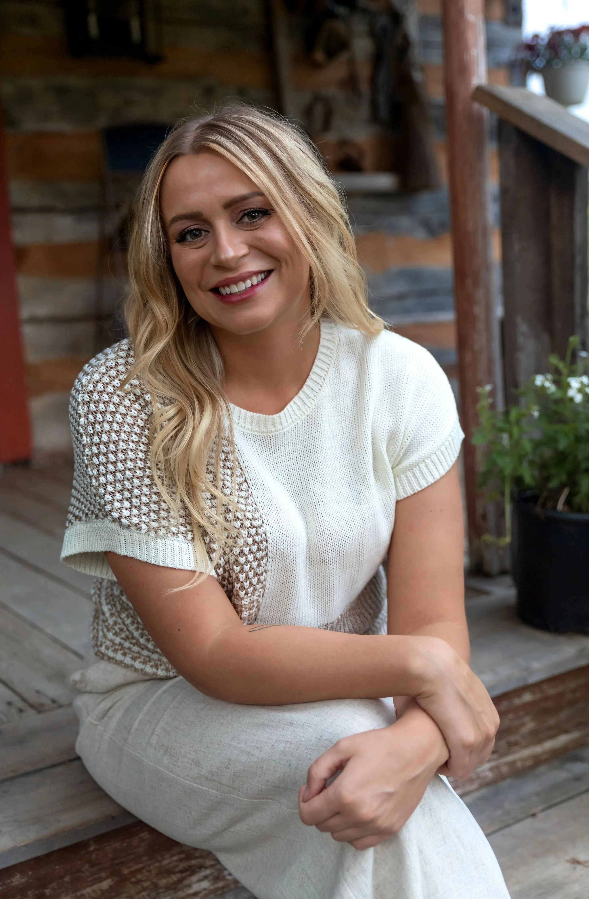 A woman with blonde, wavy hair and a warm smile, dressed in a white and beige knitted top, sitting on a wooden porch with rustic wooden walls in the background and a potted plant nearby.
