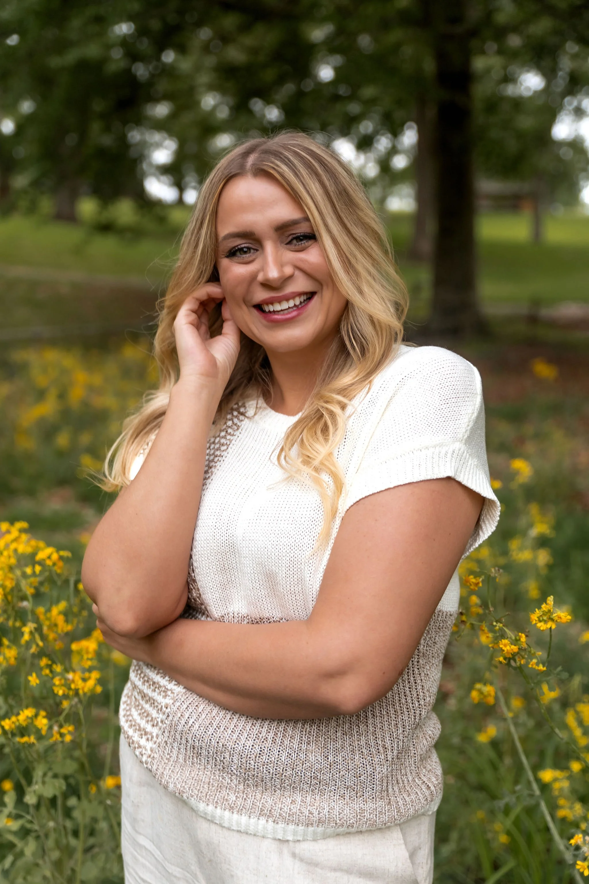 A smiling woman with blonde wavy hair, dressed in a white short-sleeve top, standing in a field of yellow flowers with trees in the background.