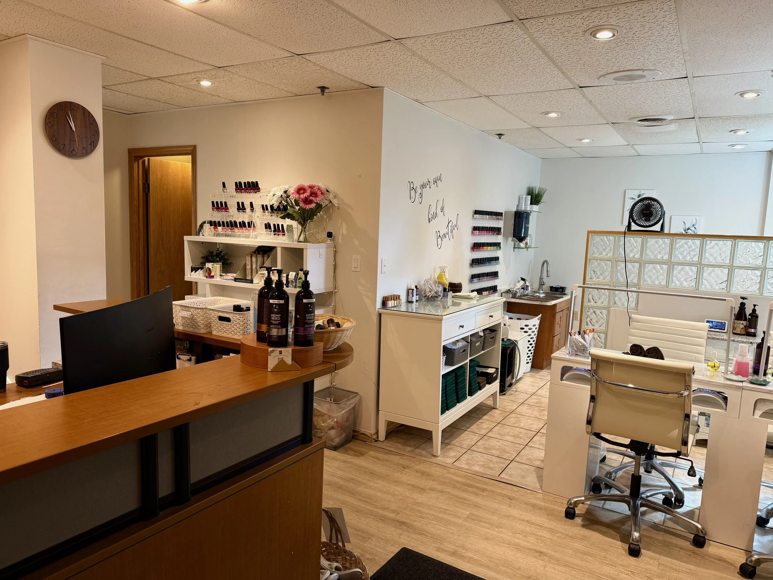 Nail salon reception area with a wooden front desk, shelves with nail polish, a treatment chair, and manicure station.