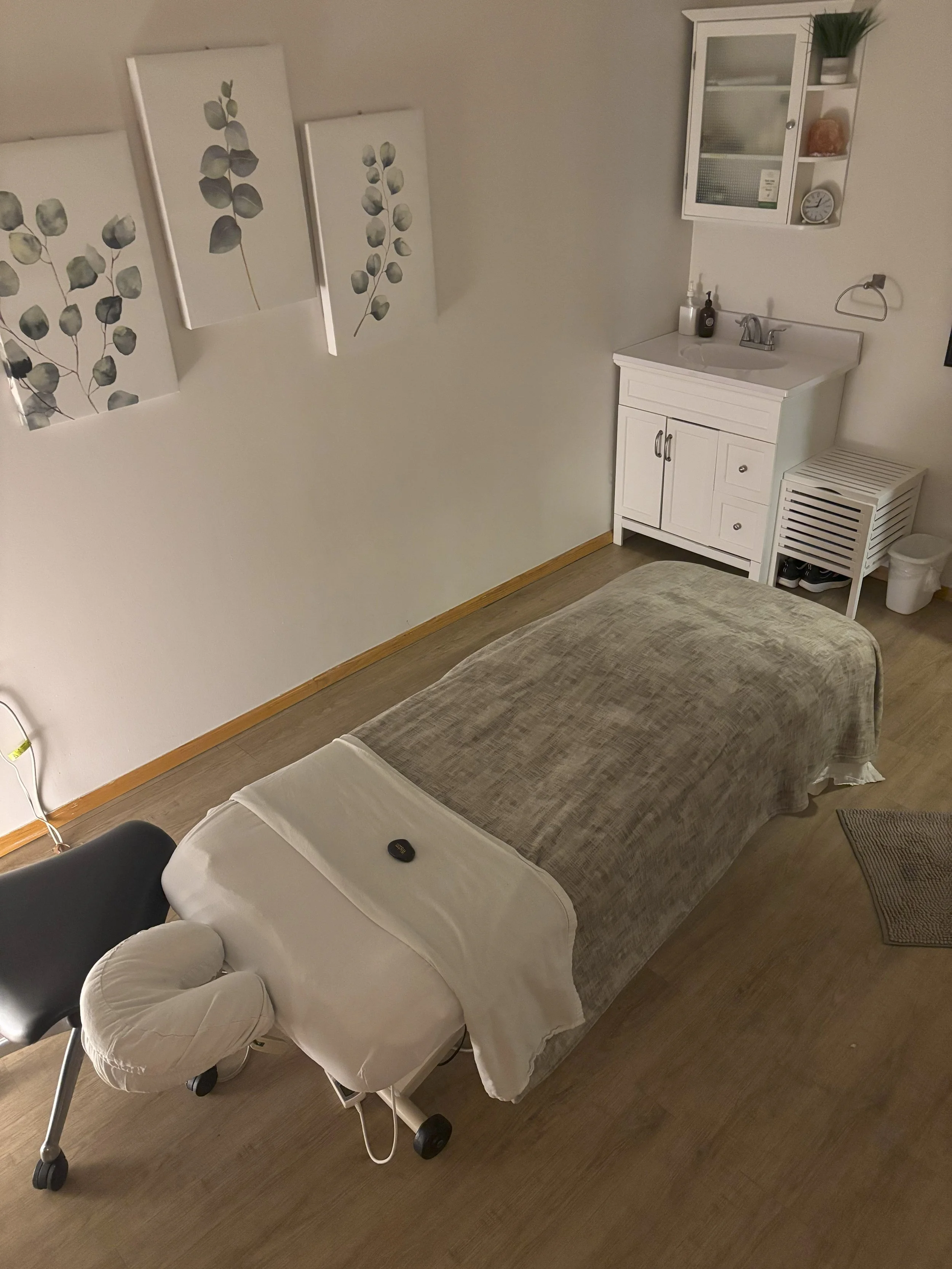 Massage table with a face cradle and a remote, situated in a room with wooden flooring, a white sink cabinet with a mirror above, decorative paintings of eucalyptus, and a small white shelf with plants and a clock.