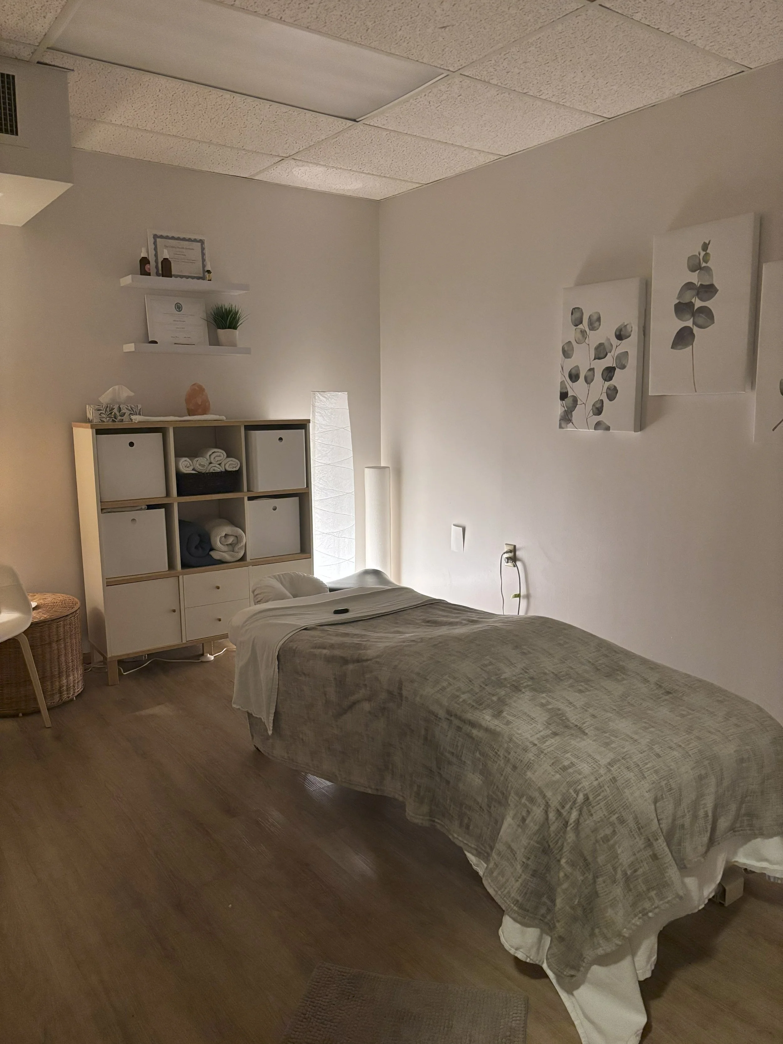 A massage therapy room with a massage table covered by a beige blanket, white pillows, a white lamp on the left, and framed botanical artwork on the wall. There is a white shelf with towels and a cabinet with additional towels near the massage table.