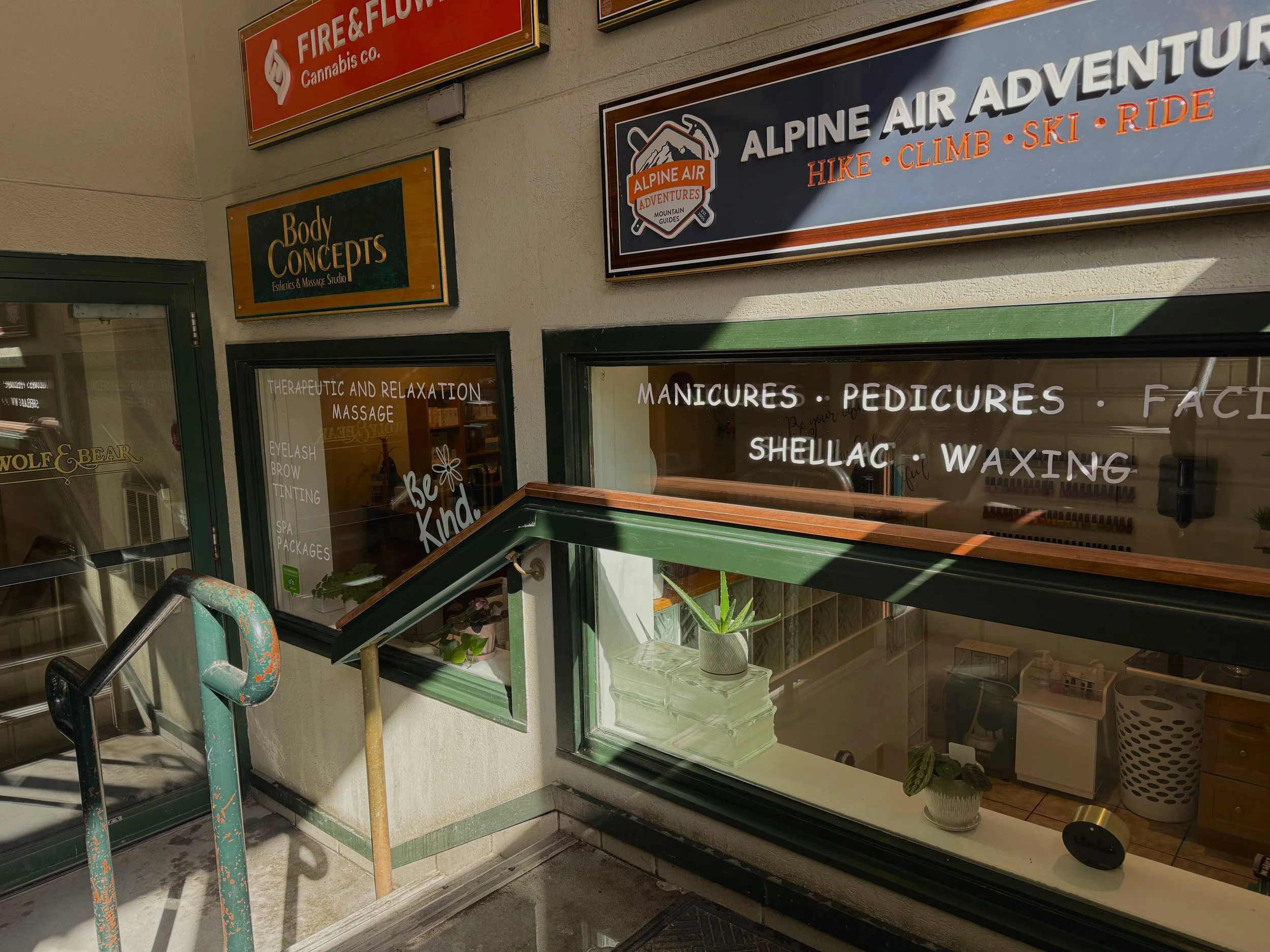 Exterior window of a beauty salon with signs listing services such as manicures, pedicures, shellac, waxing, massage, and spa packages. Inside, plants are visible.