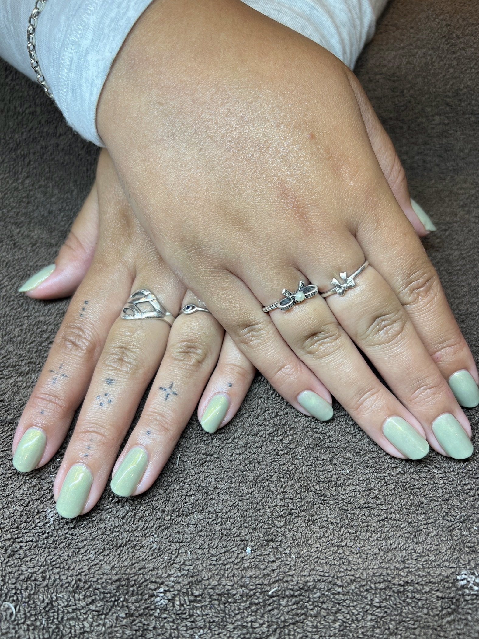 Hands with gold nail polish, tattoos, and multiple silver rings, resting on a dark textured surface.