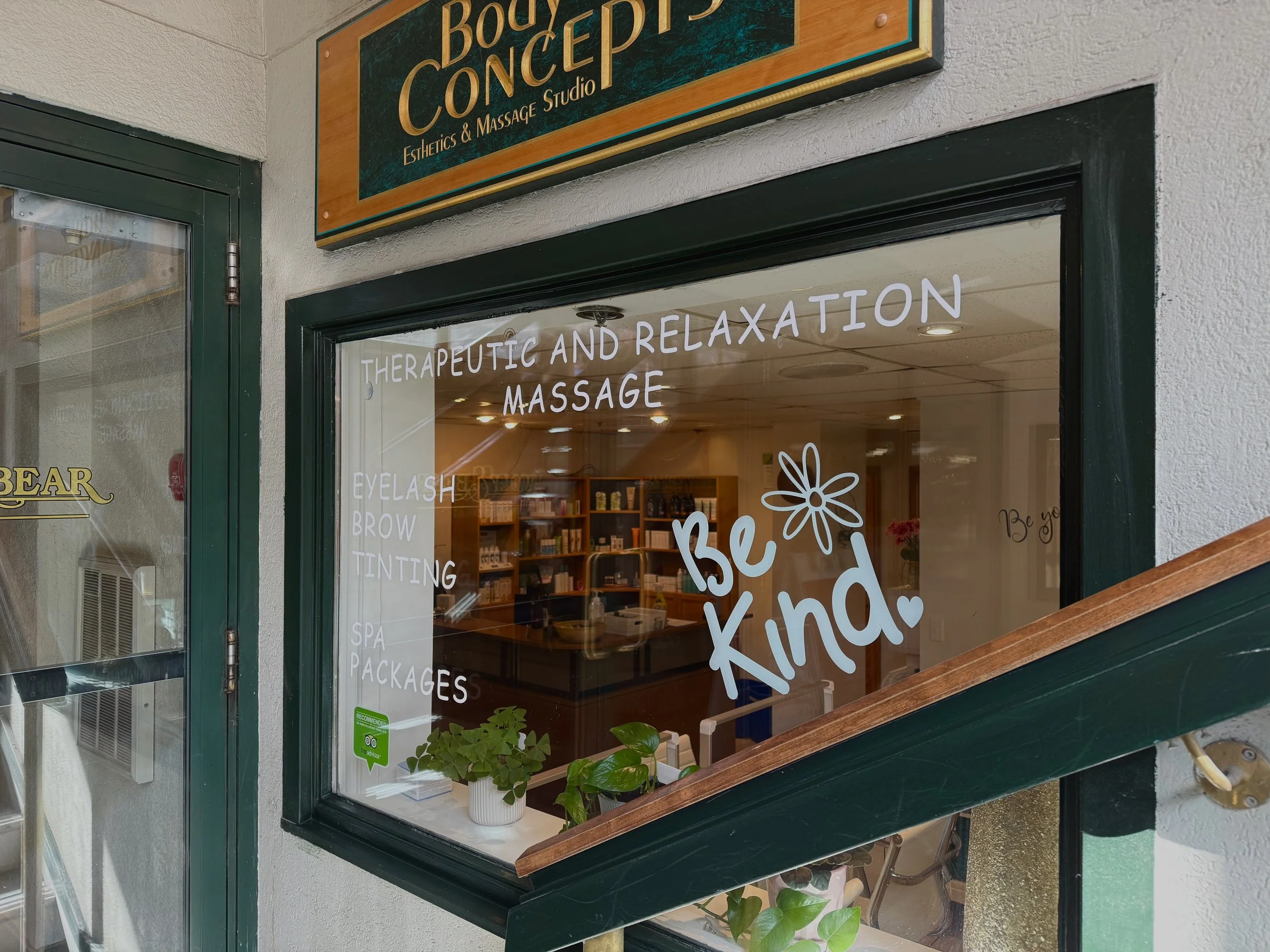 The window of a wellness studio with white text promoting services like therapeutic massage, eyelash and eyebrow tinting, and spa packages, along with the phrase 'Be Kind' and a flower drawing.