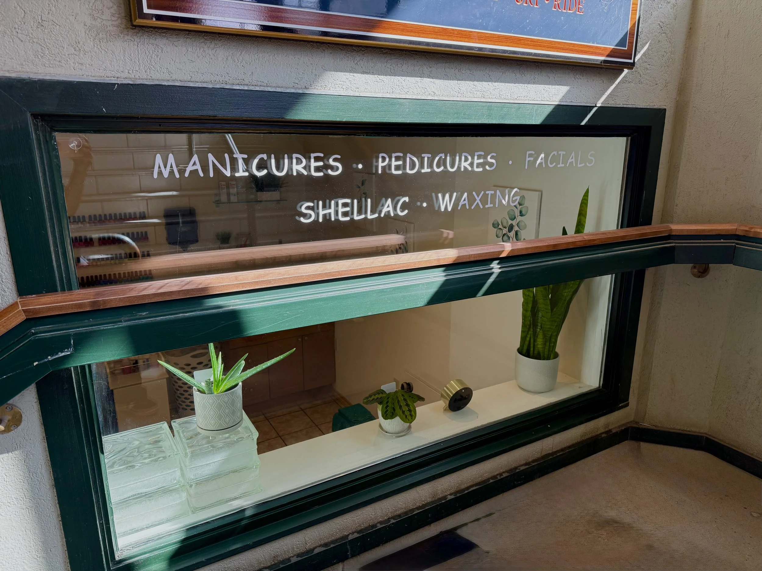 Window display with white text listing services: manicures, pedicures, facials, shellac, waxing. Inside, there are potted plants, glass blocks, and a wall with framed art.