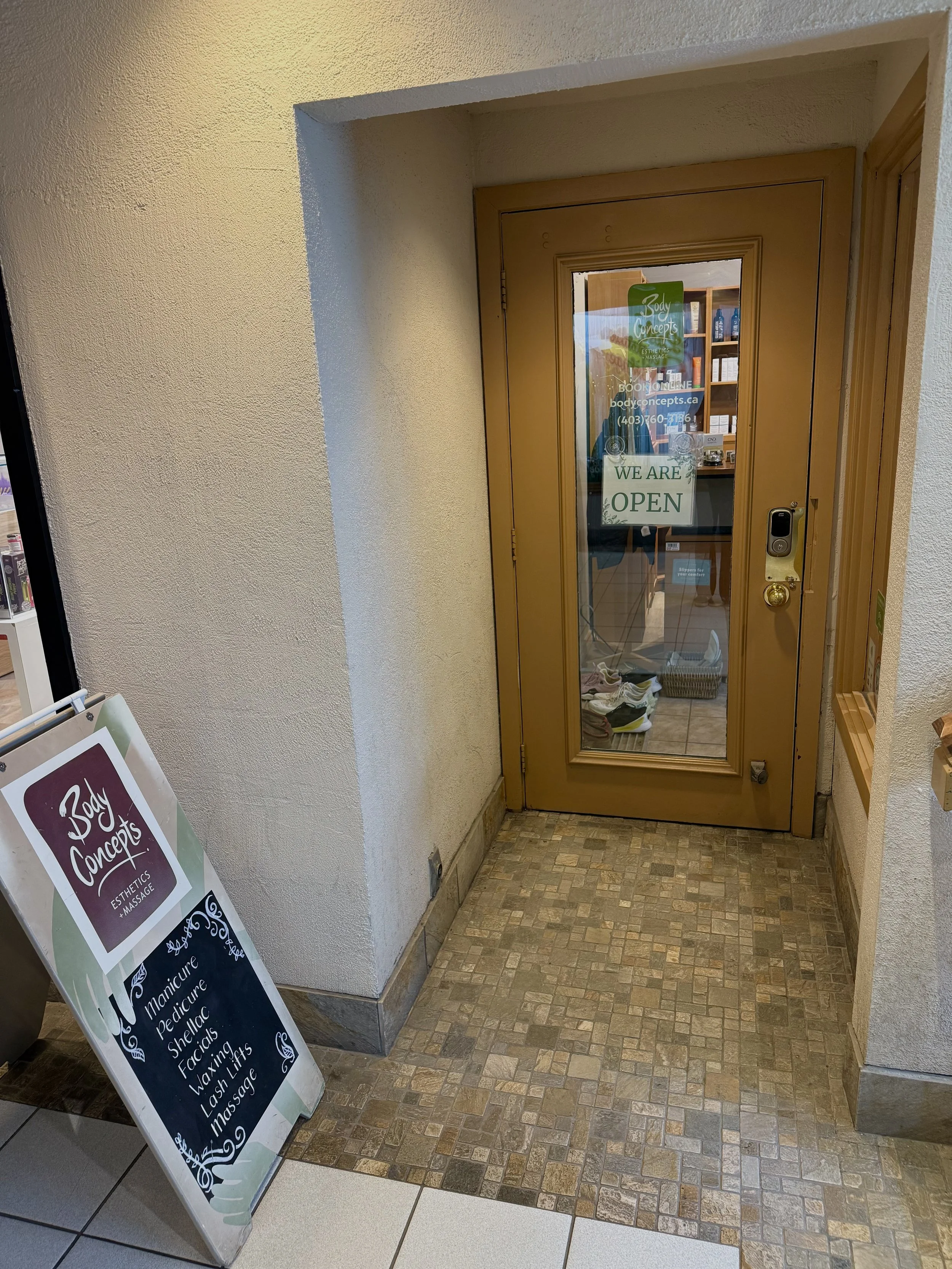 Door with a sign that says "We Are Open" next to a stand-up signboard listing services like manicure, pedicure, shellac, facials, waxing, lash lifts, and massage at a beauty business.