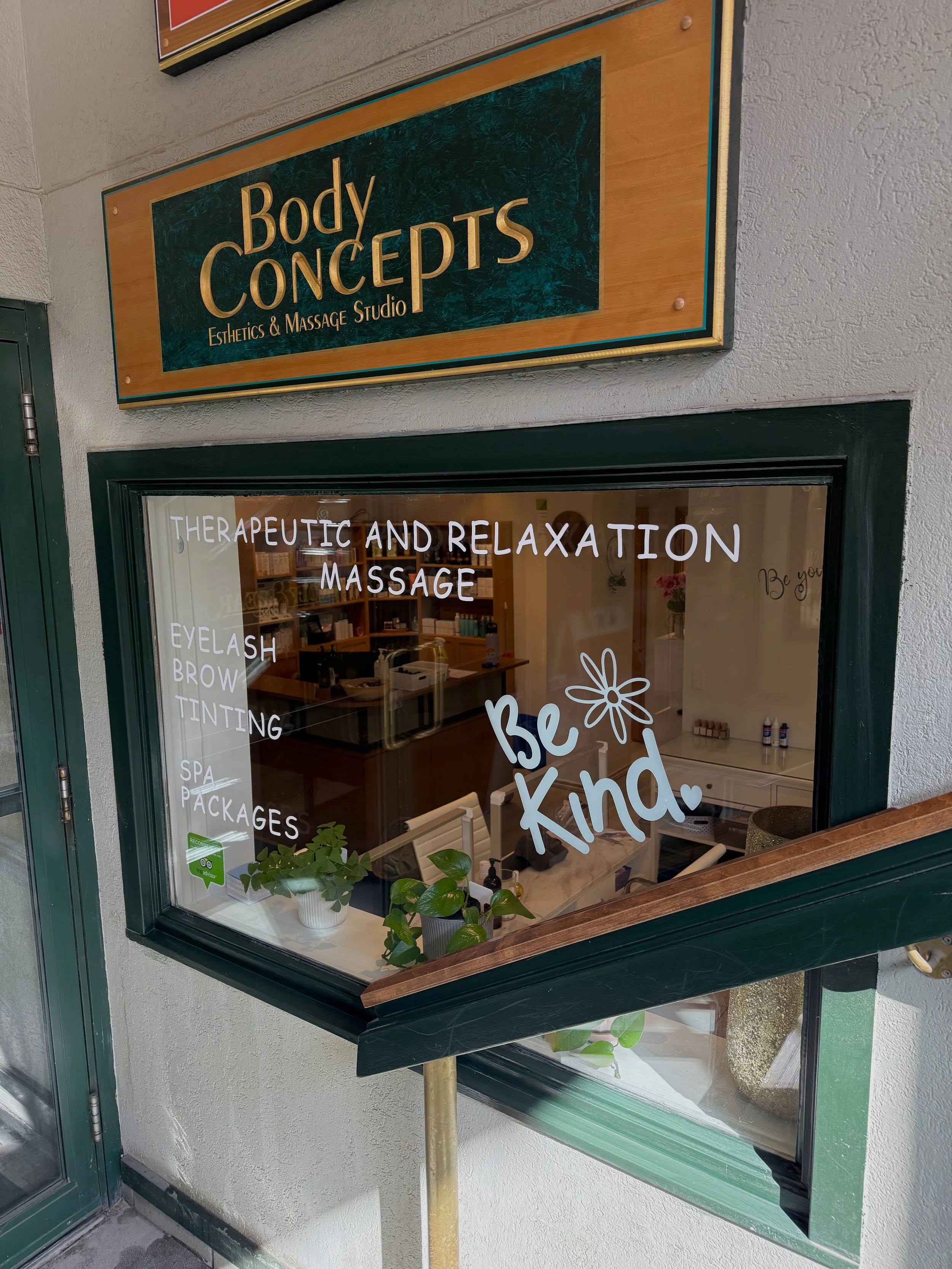 Exterior window display for Body Concepts Esthetics & Massage Studio, featuring handwritten white text promoting therapeutic and relaxation massage, eyelash, brow, tinting, spa packages, with a decorative flower and the phrase 'Be kind', and indoor p