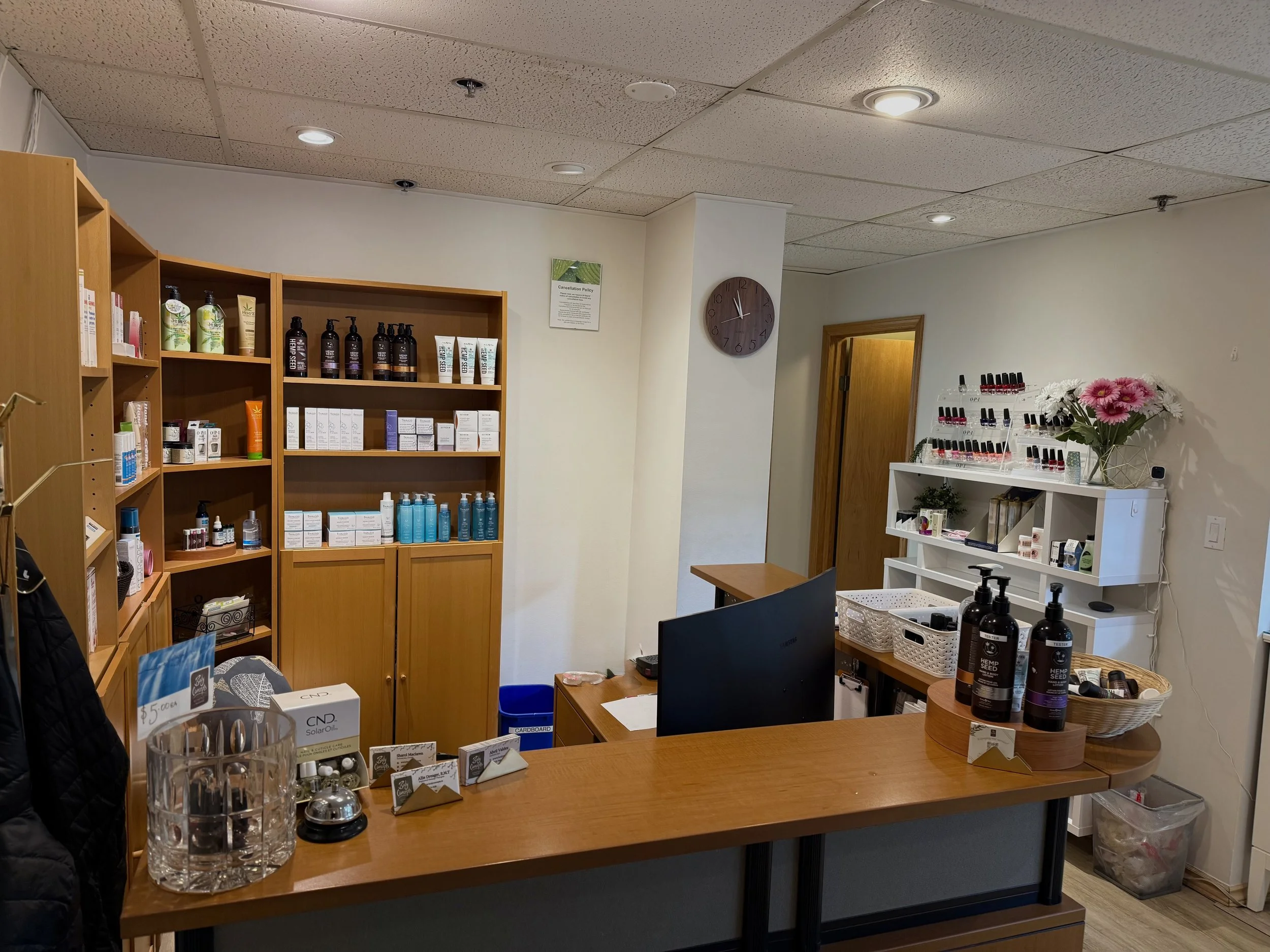 A store or small business interior with wooden shelves and a counter, displaying skincare, beauty, and wellness products such as lotions, oils, nail polishes, and personalized products, with a clock on the wall, flowers, and various bins and baskets.