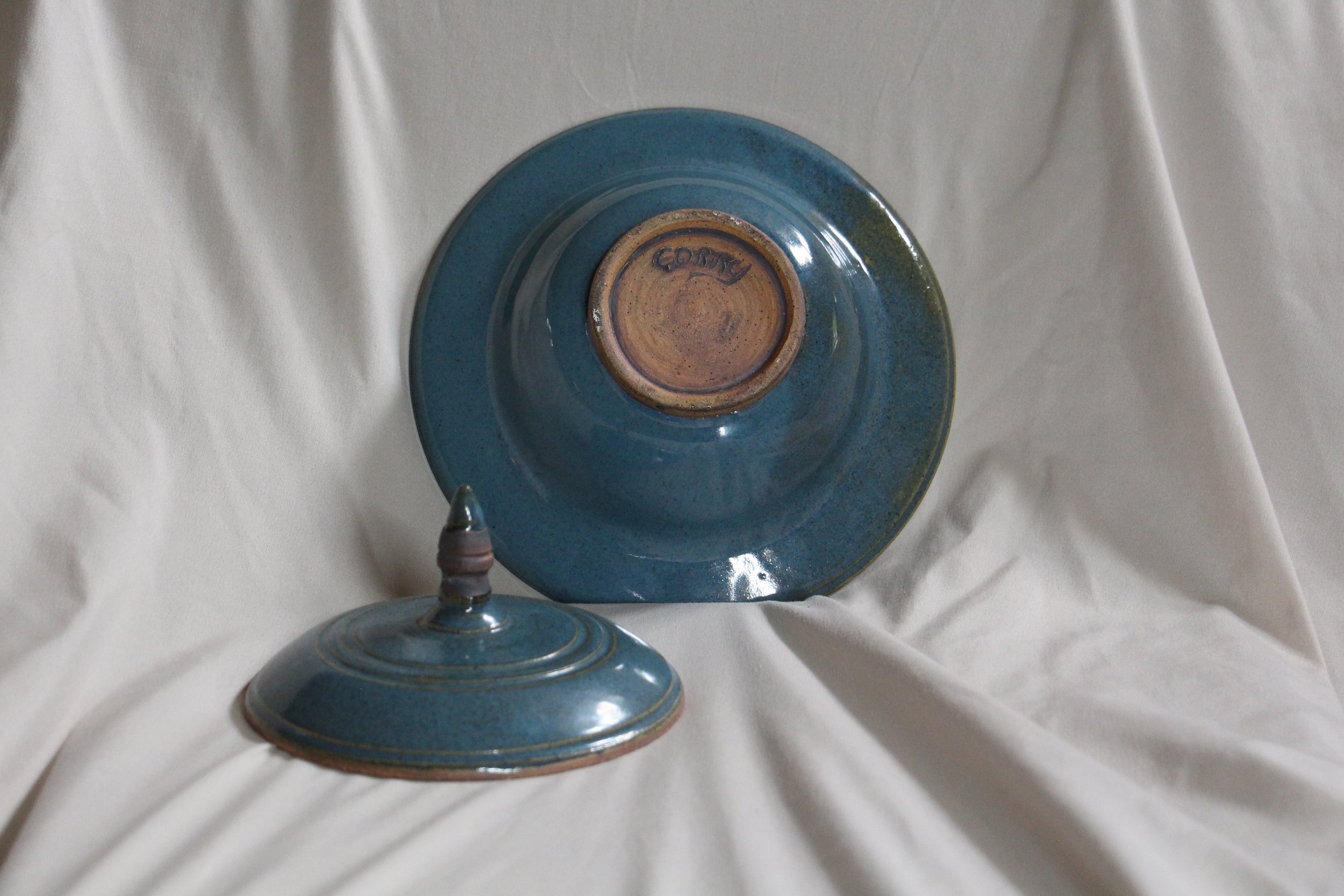 A ceramic upside-down bowl with a brown underside and the word 'COSY' inscribed on it, with a matching ceramic lid beside it, both with a blue glaze, set against a white fabric background.