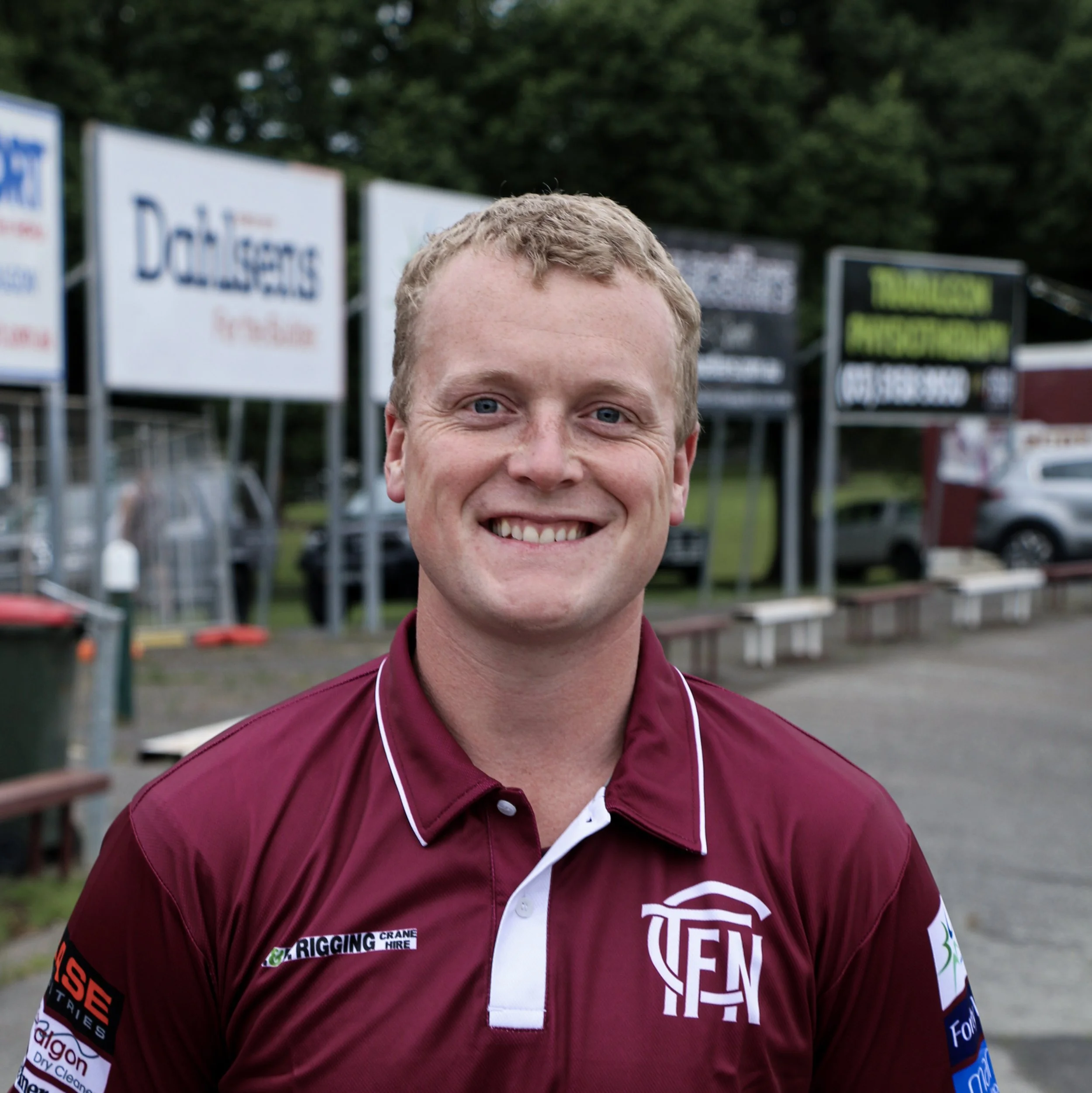 A smiling man wearing a maroon polo shirt with various logos, standing outdoors with signs and parked cars in the background.