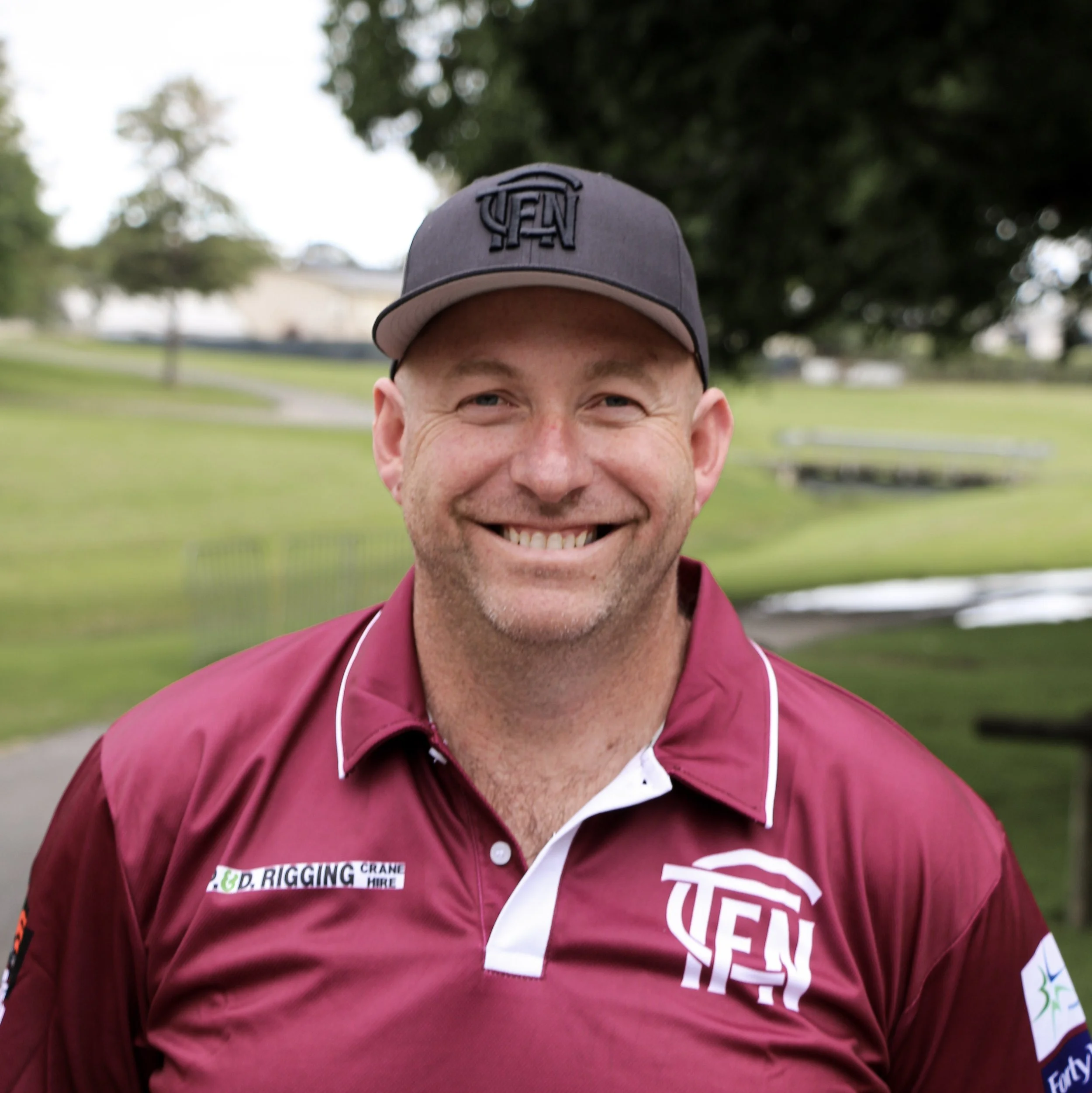 A person wearing a gray cap with a logo, smiling, with a park and trees in the background, partially visible from the nose up, wearing a maroon jacket with patches.