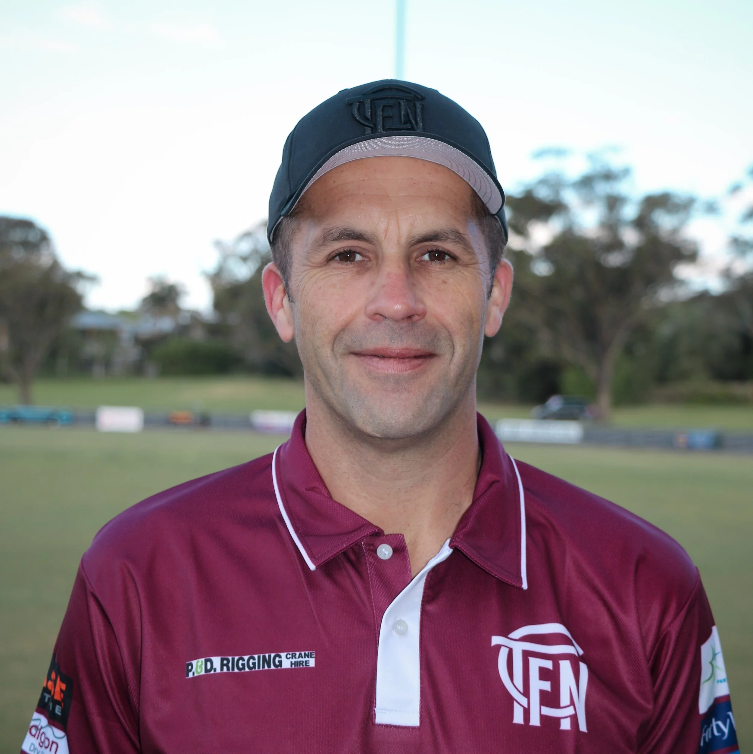 A man wearing a maroon sports polo shirt and a black cap, standing outdoors on a grassy field with trees in the background.